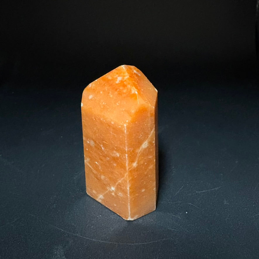 A polished orange calcite stone crystal with a geometric, stepped pyramid shape, standing upright on a dark, reflective surface.