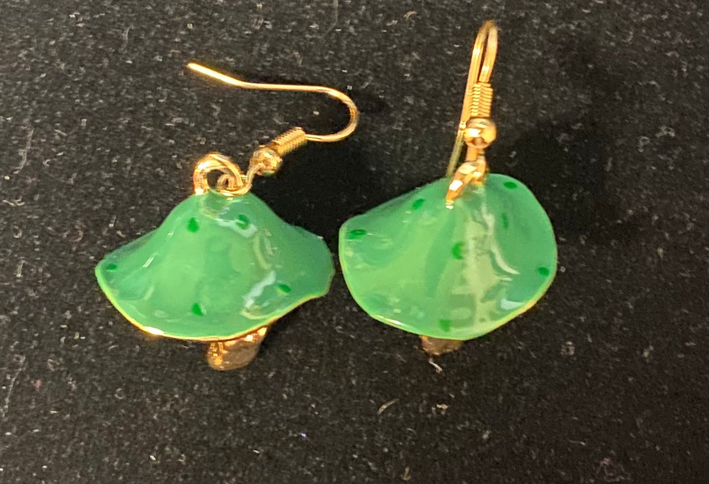 A pair of mushroom-shaped dangling earrings with glossy green enamel bodies and gold-tone hooks, displayed on a dark textured background.