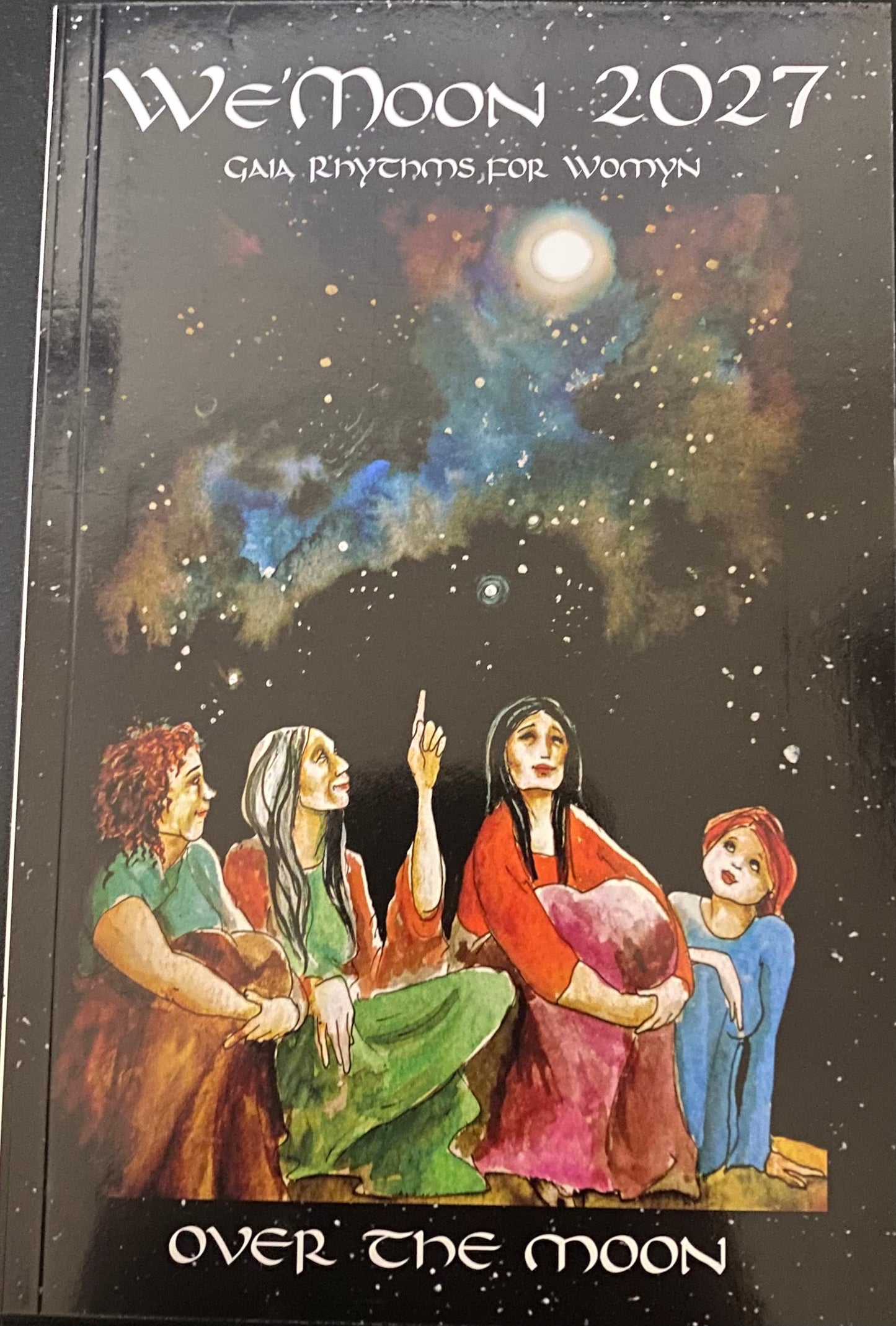 A book cover titled "We'Moon 2027: Gaia Rhythms for Women" with an illustrated night sky scene and four women looking up at a glowing moon, plus the text "Over the Moon" at the bottom.