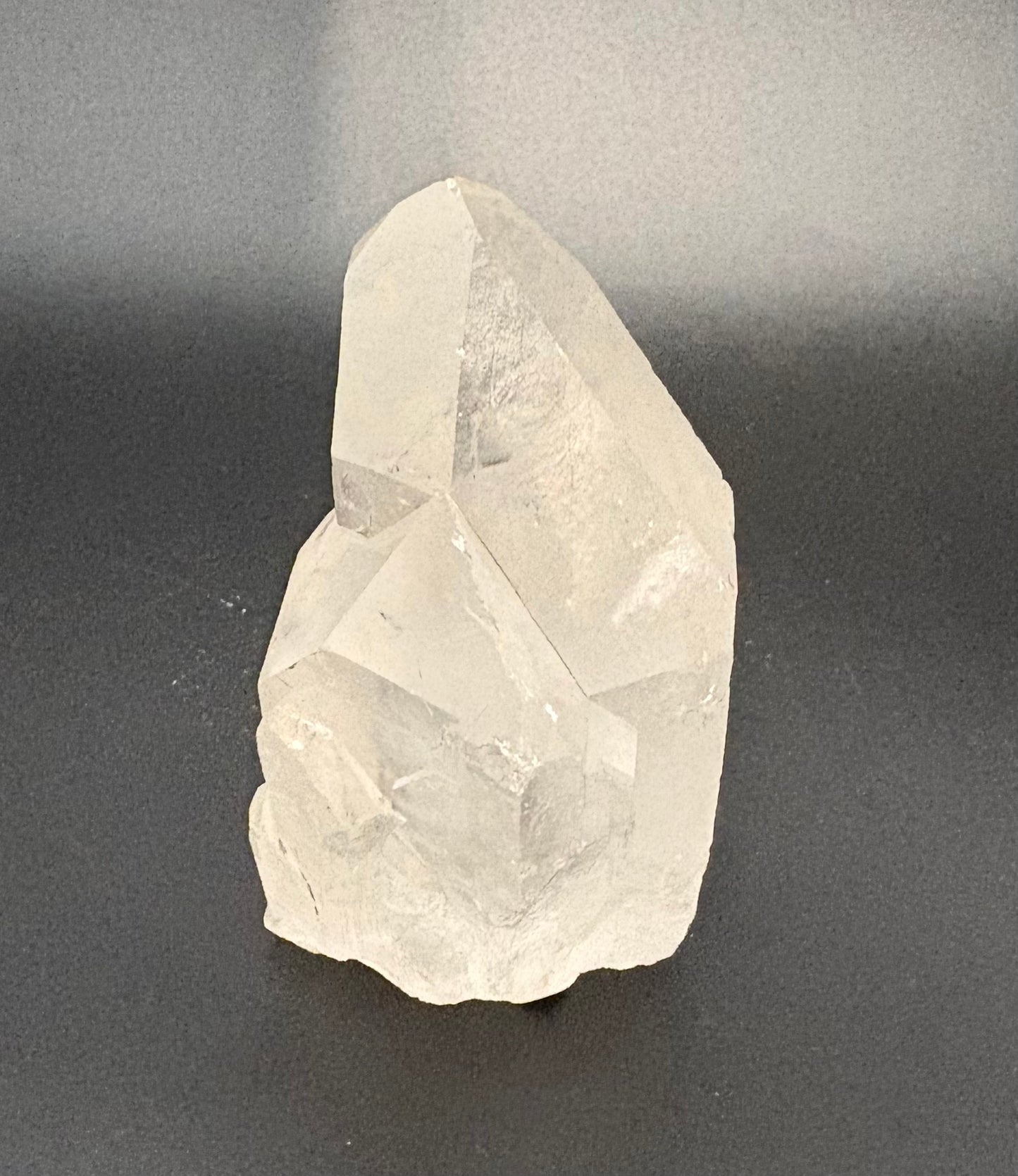 A raw, unpolished clear quartz crystal cluster with natural geometric facets, standing upright on a dark gray surface against a gray background.