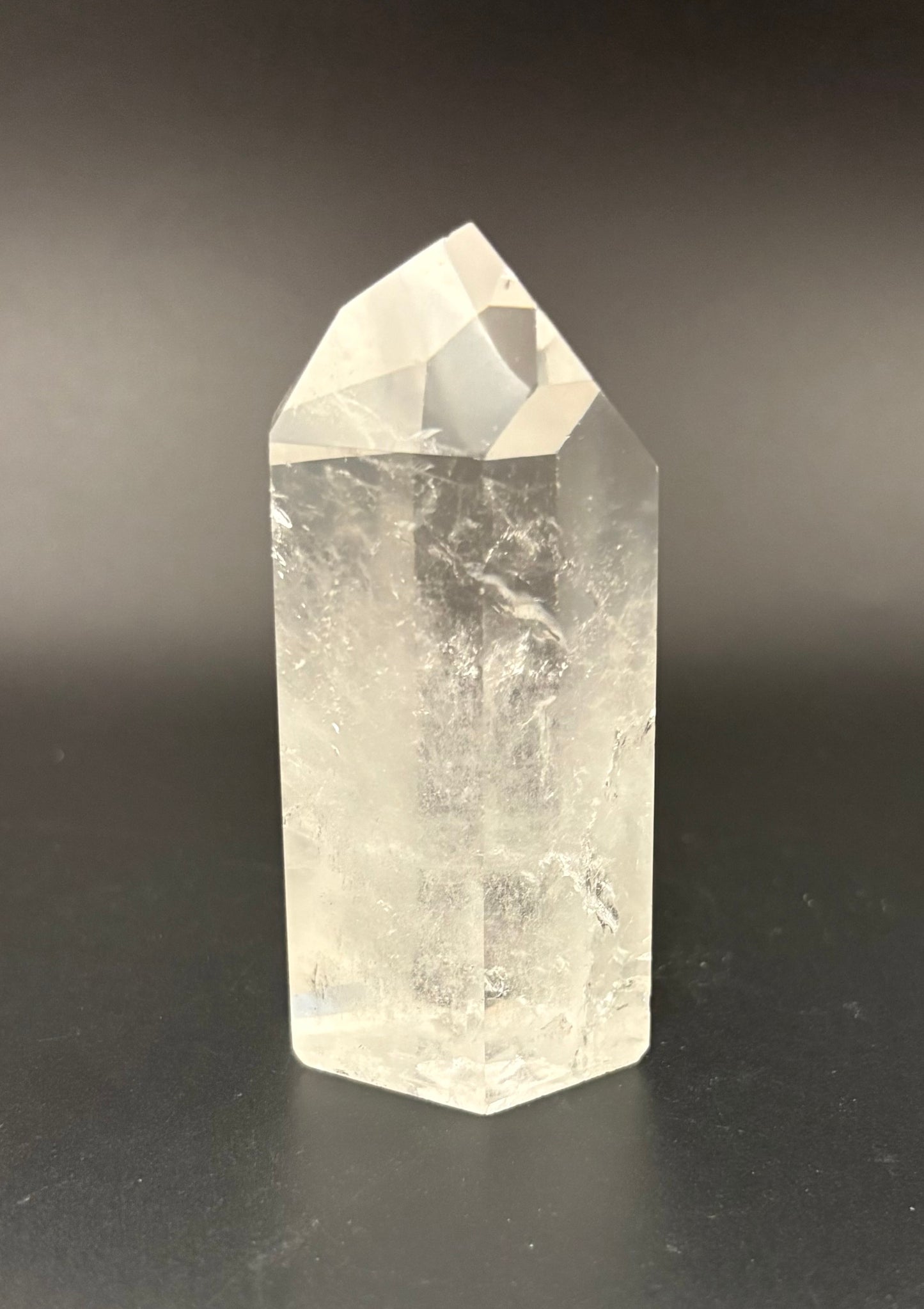 A clear quartz crystal point tower with natural internal inclusions, standing on a dark surface against a dark gray background.