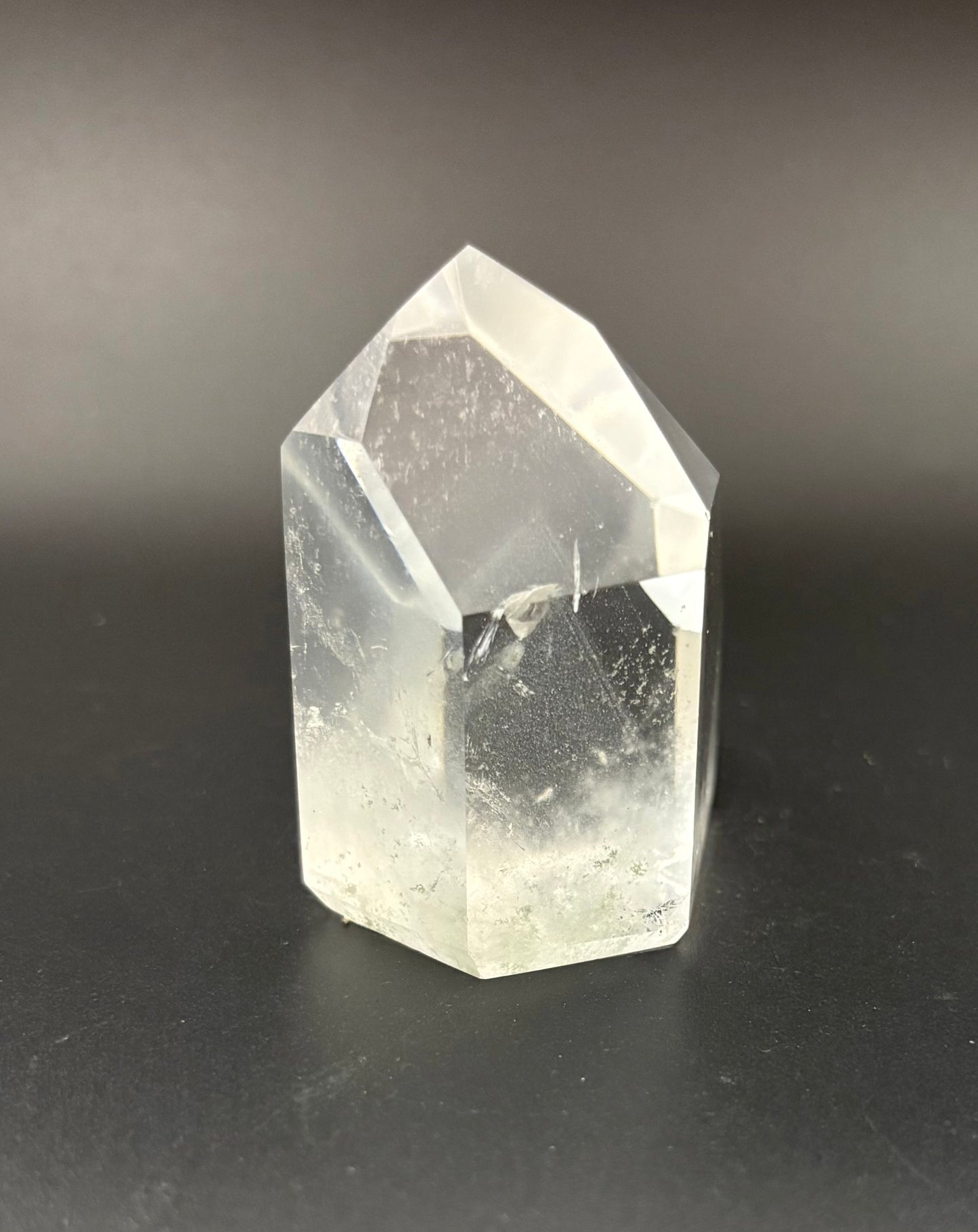 A clear quartz crystal point with natural inclusions, standing on a dark gray surface against a dark gray background.