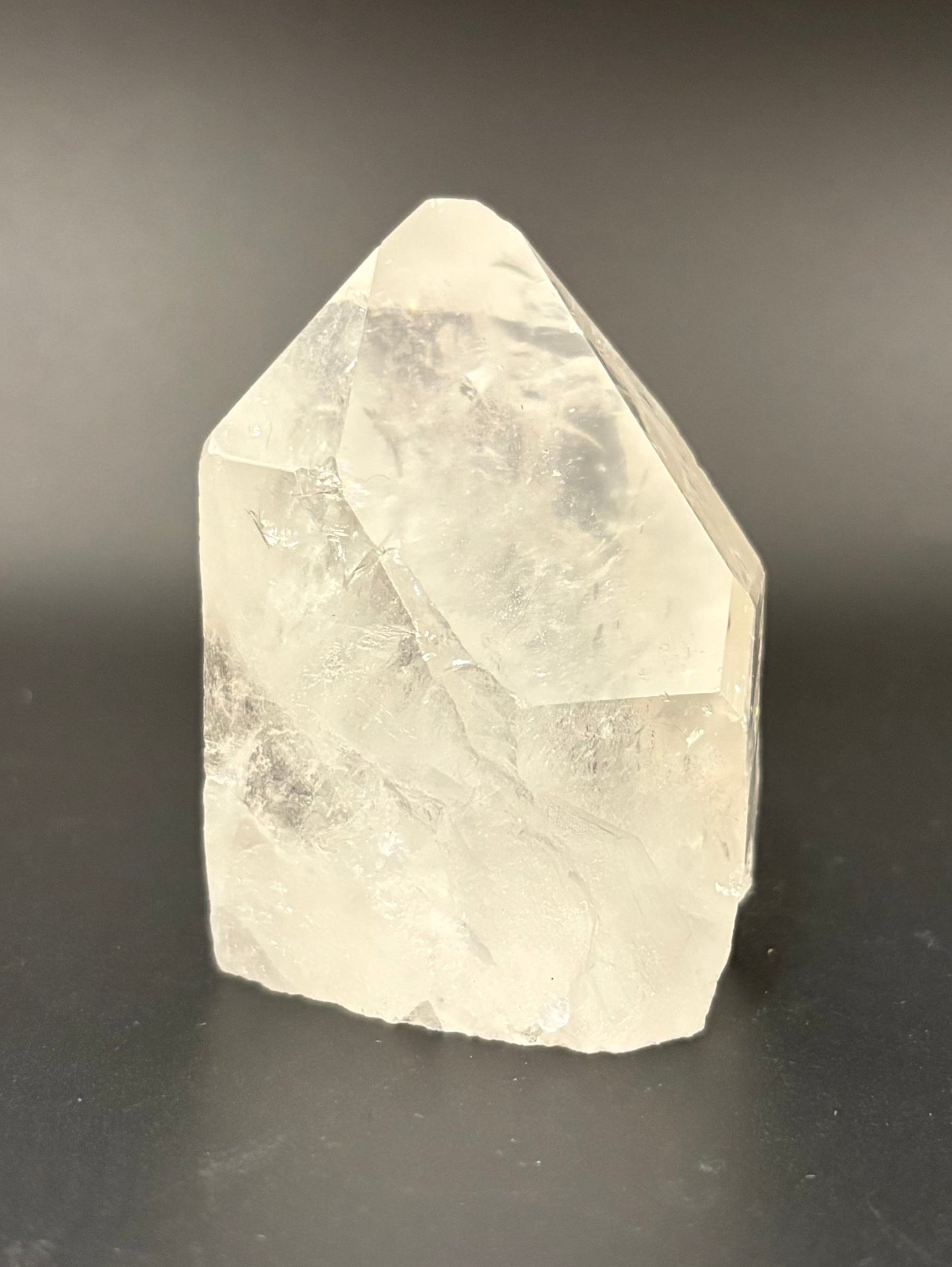 A raw, pointed crystal stone with natural facets and internal inclusions, shown on a dark gray background.