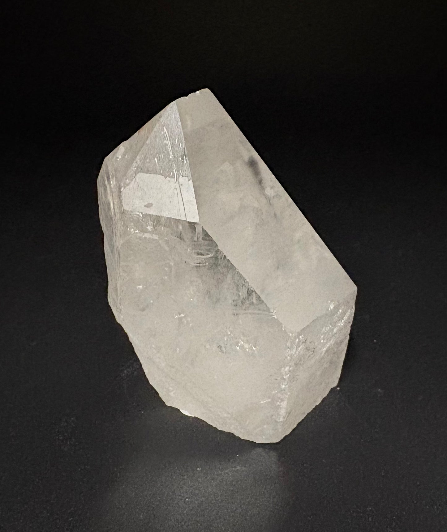 A raw, unpolished clear quartz crystal point with natural facets, standing upright on a dark, reflective surface against a black background.