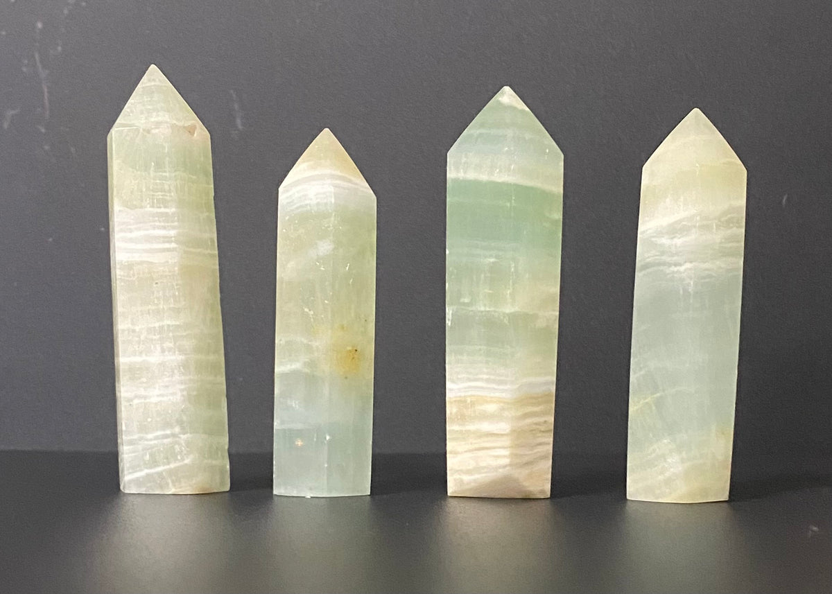 Four pointed crystal tower pendants made of pale green and cream-colored calcite, arranged in a row against a dark gray background.