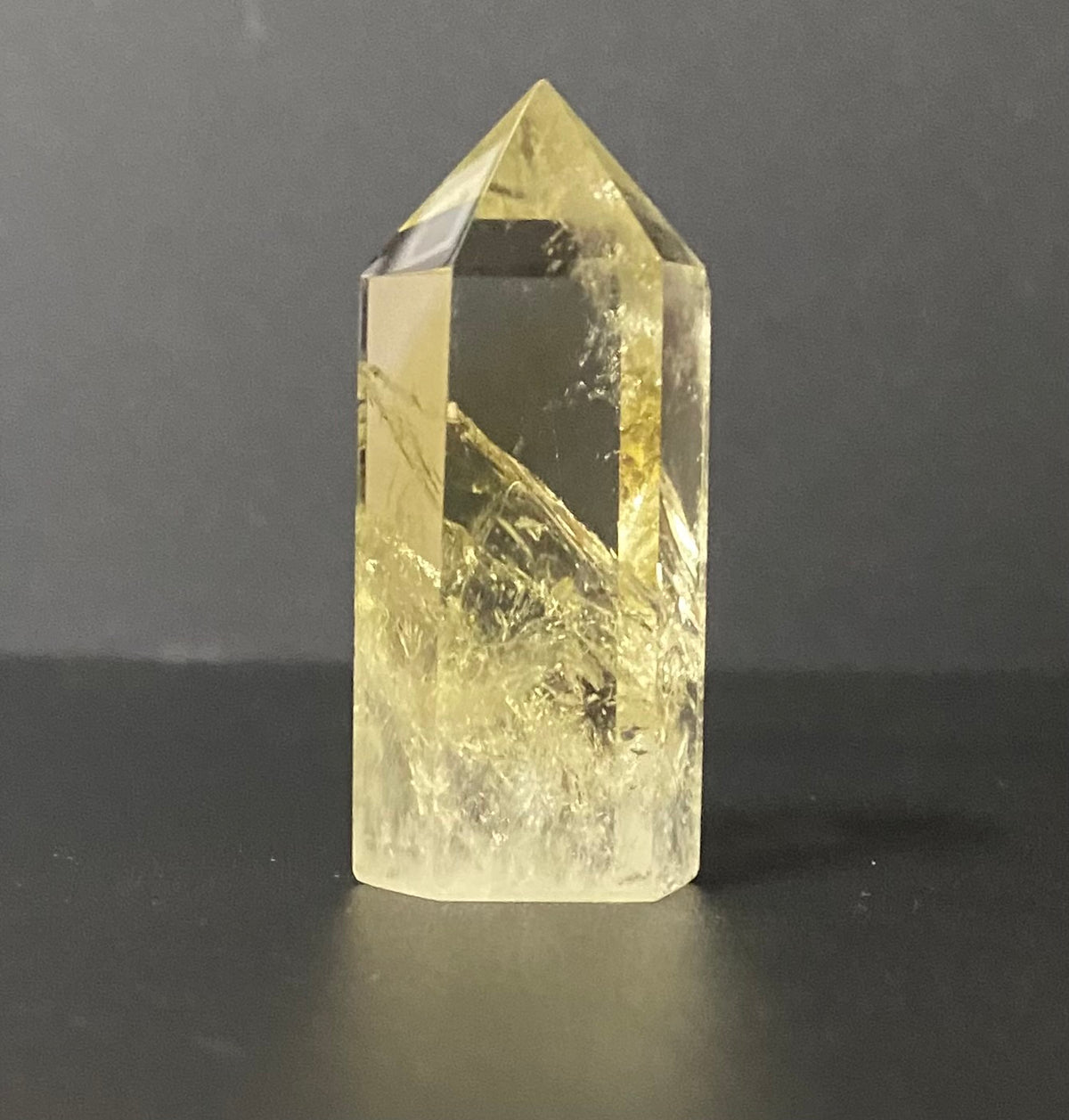 A natural pointed citrine crystal tower, featuring a pale yellow hue and prominent internal inclusions, standing on a dark surface against a dark gray background.