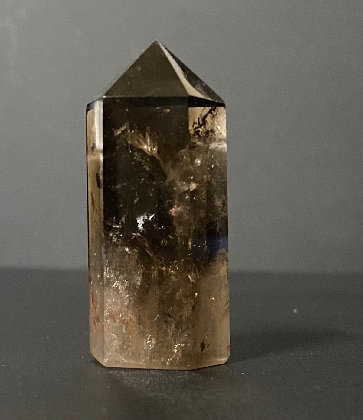 A natural smoky quartz crystal point with a hexagonal shape, showing translucent brown and amber tones with internal inclusions, standing on a dark surface against a neutral gray background.