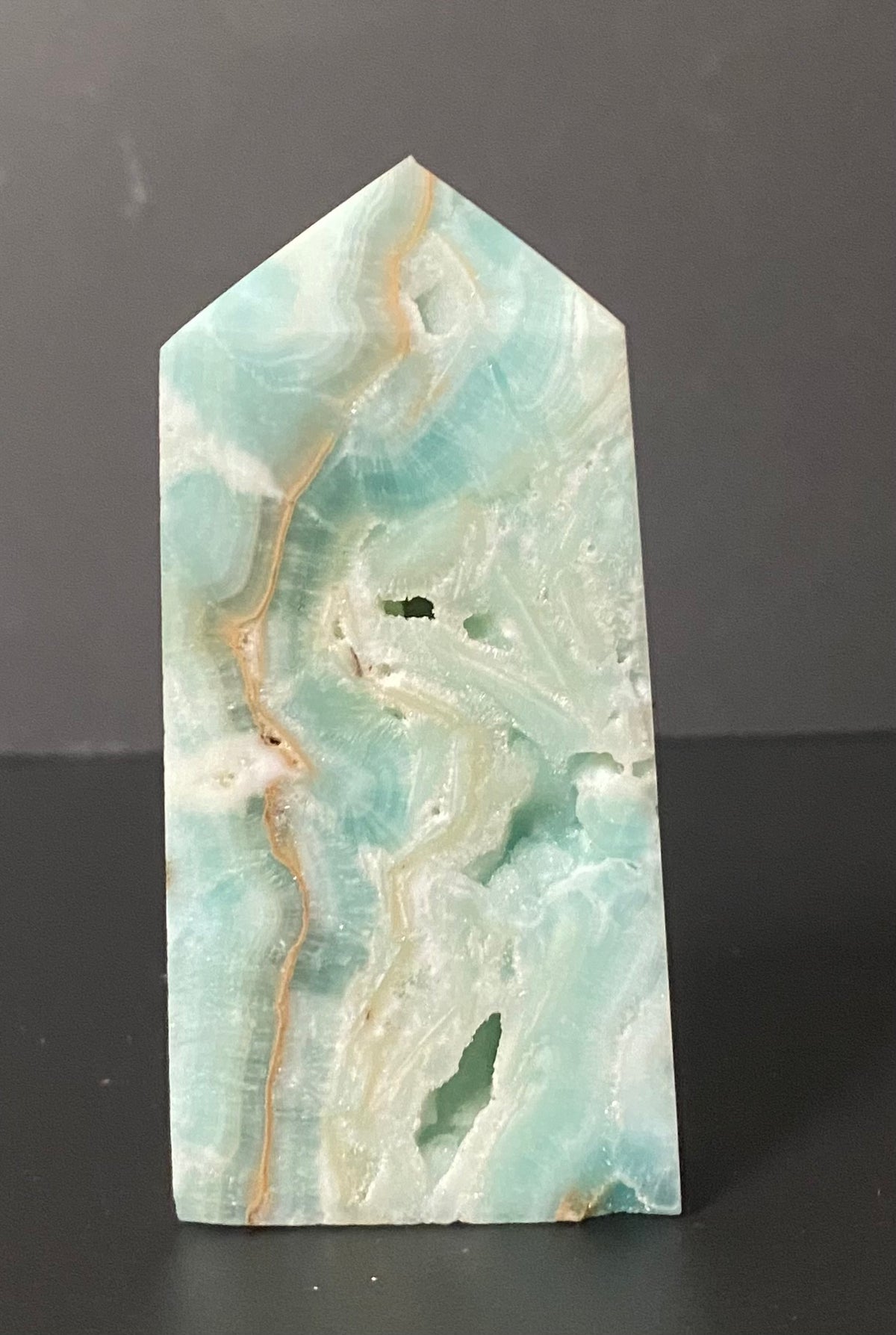 A pointed rectangular polished stone or crystal with a marbled pattern of light aqua, mint green, cream, and tan veining, standing upright on a dark surface against a dark gray background.