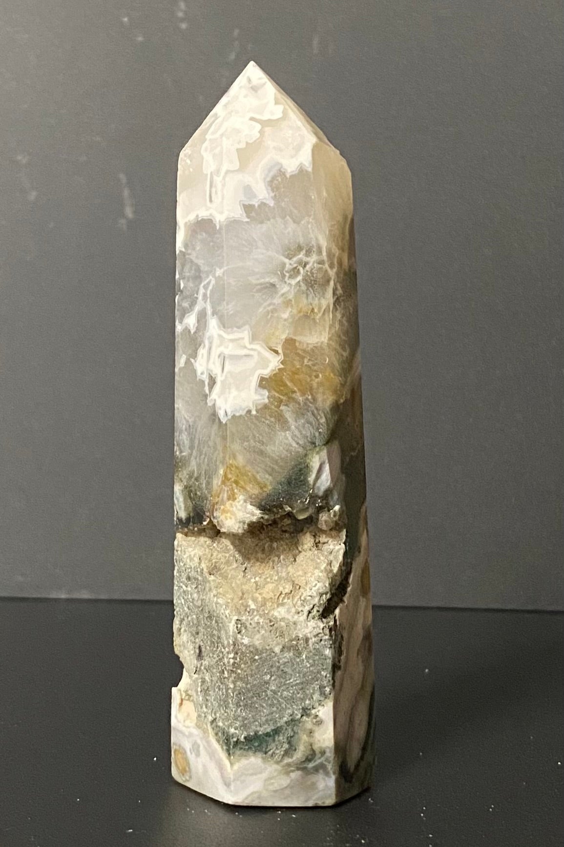 A natural pointed crystal Tree Agate tower or mineral spire with marbled patterns of white, gray, beige, and green, standing on a dark surface against a dark gray background.
