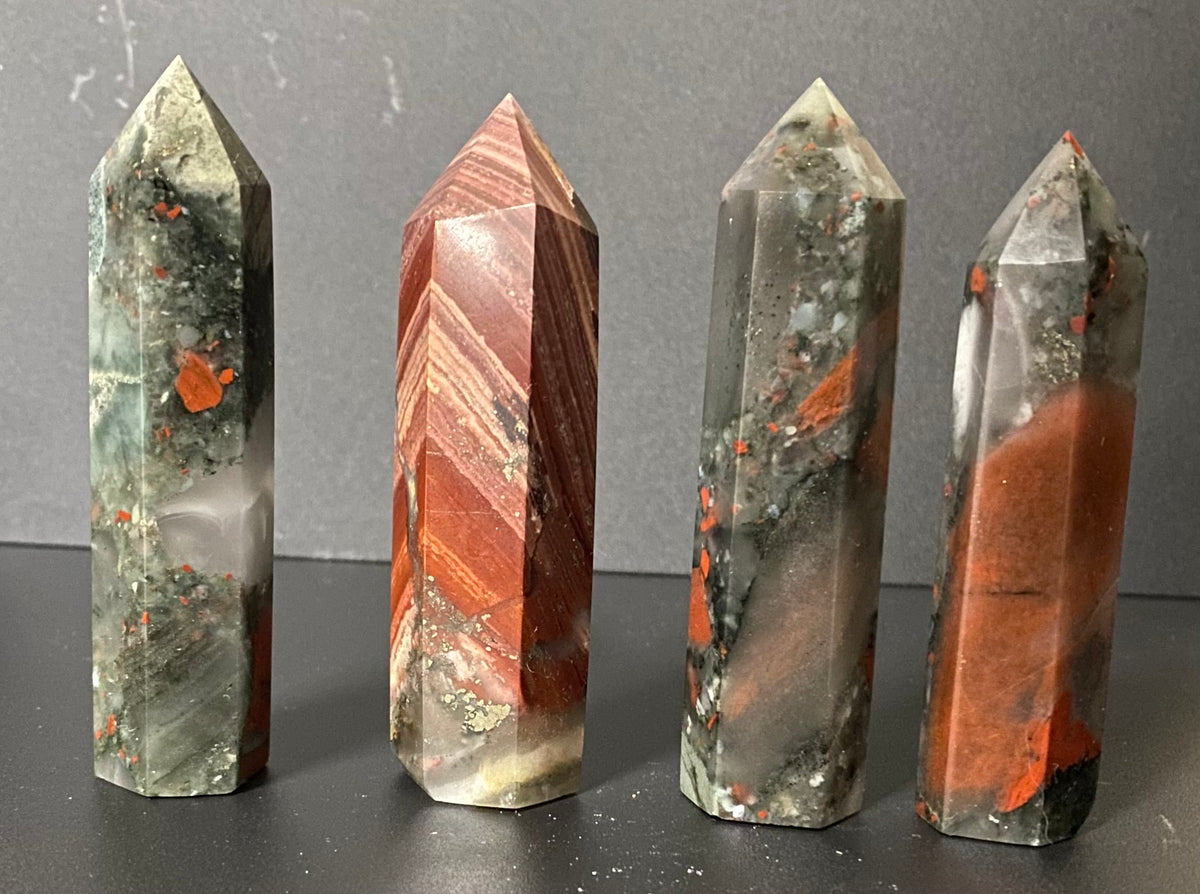 Four polished crystal points Bloodstone with natural banding and inclusions, displayed upright on a dark surface against a gray background.
