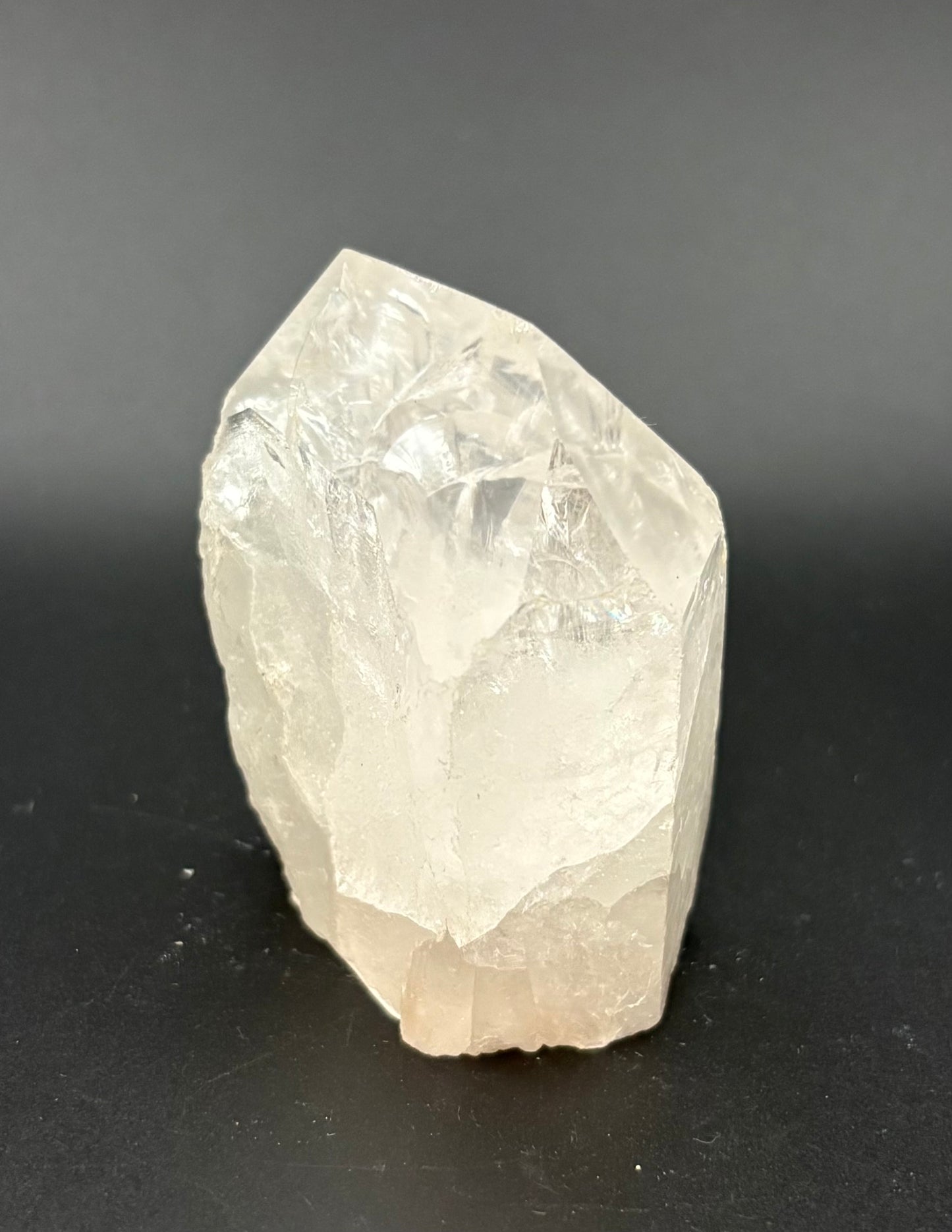 A raw, unpolished clear quartz crystal with natural facets, standing upright on a dark gray surface against a dark gray background.