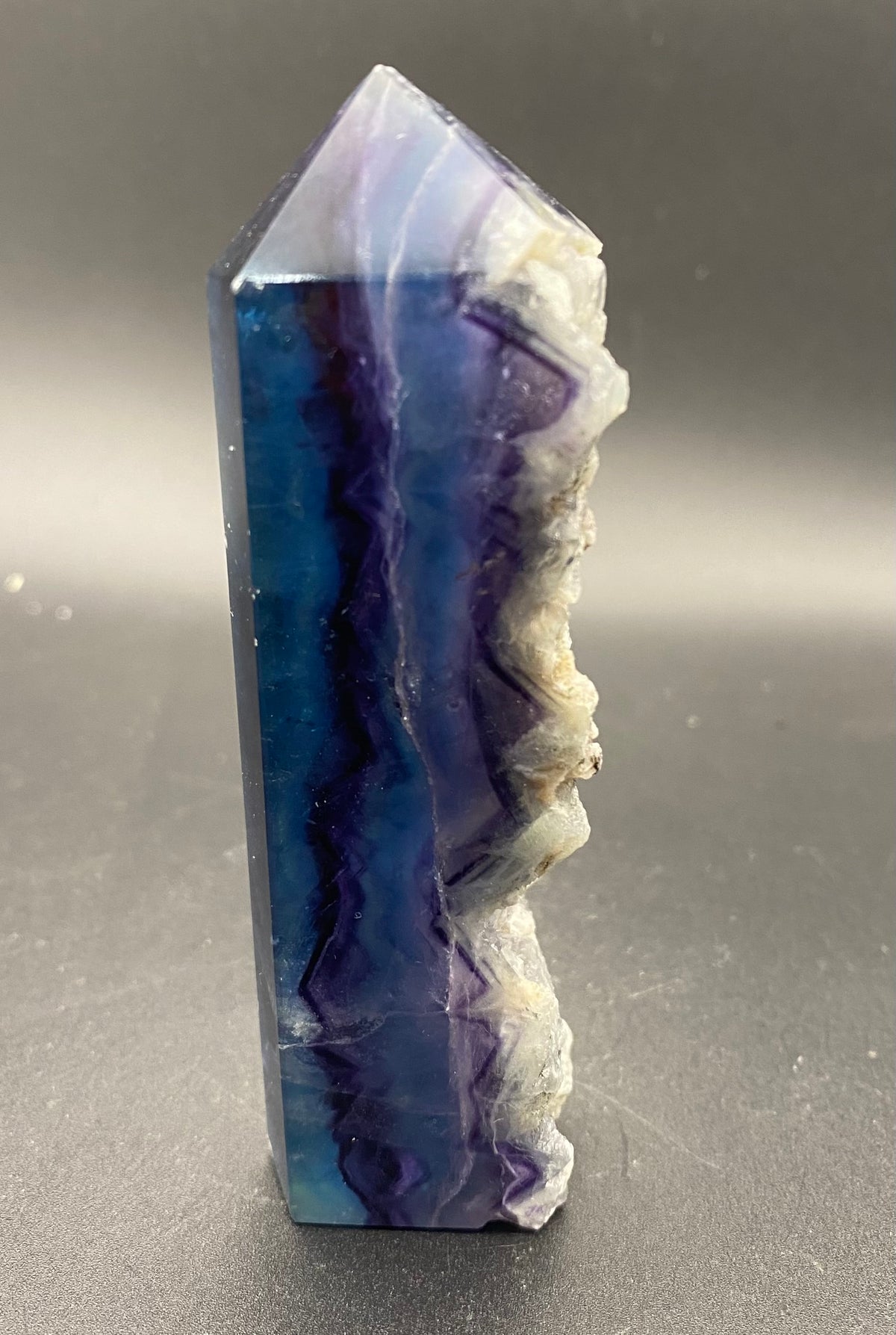 A polished fluorite natural crystal point showing vibrant blue and purple agate-like bands with a rough, textured white outer layer, standing upright on a dark gray surface.