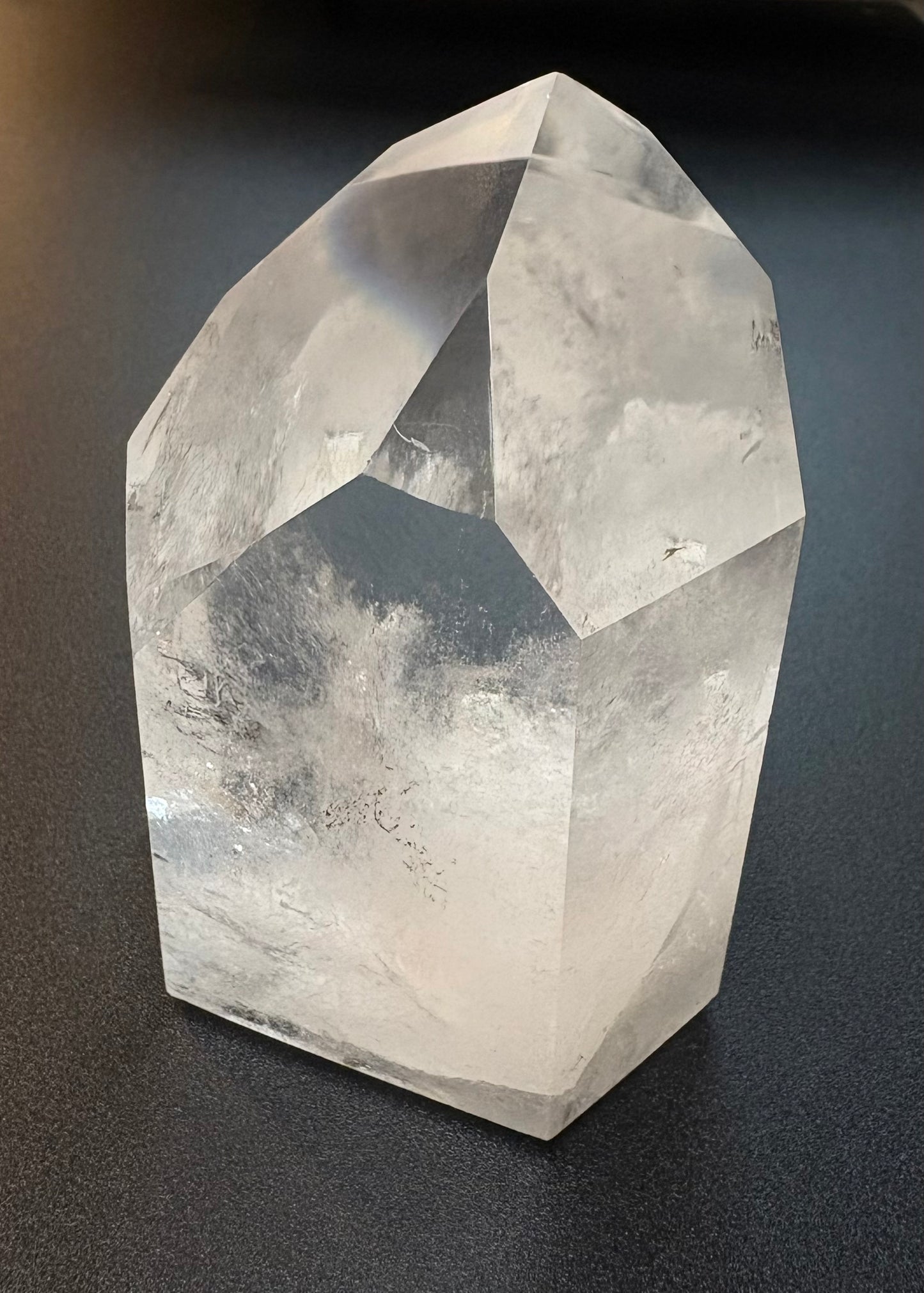 A natural clear quartz crystal point with faceted geometric edges, displayed on a dark gray surface.