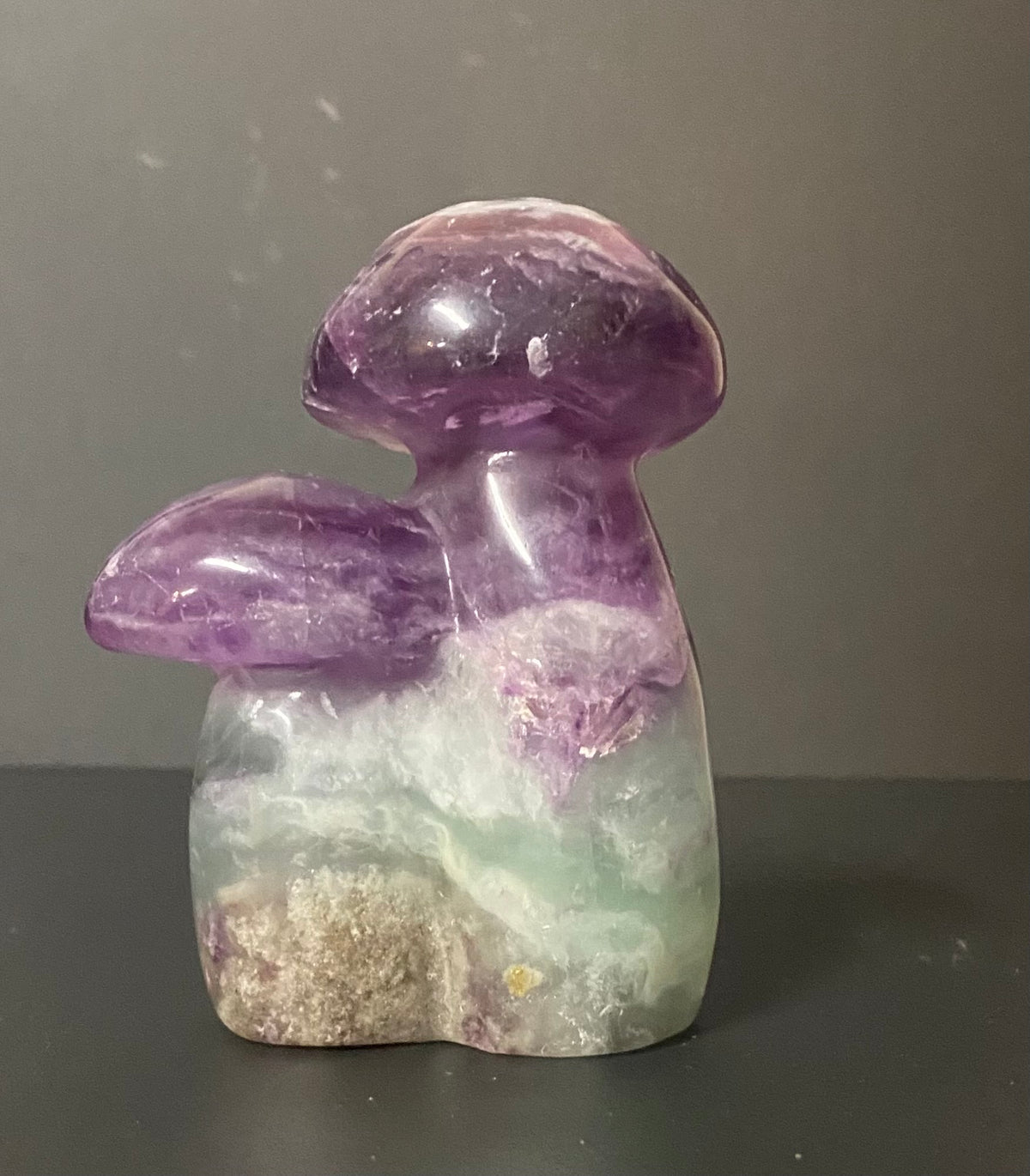 A carved fluorite and fluorite mushroom with a purple and green gradient body, resting on a dark surface against a neutral gray background.