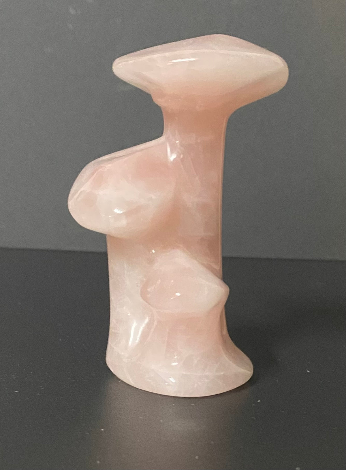 A small mushroom sculptural made of rose quartz pale pink stone, photographed against a dark gray background.