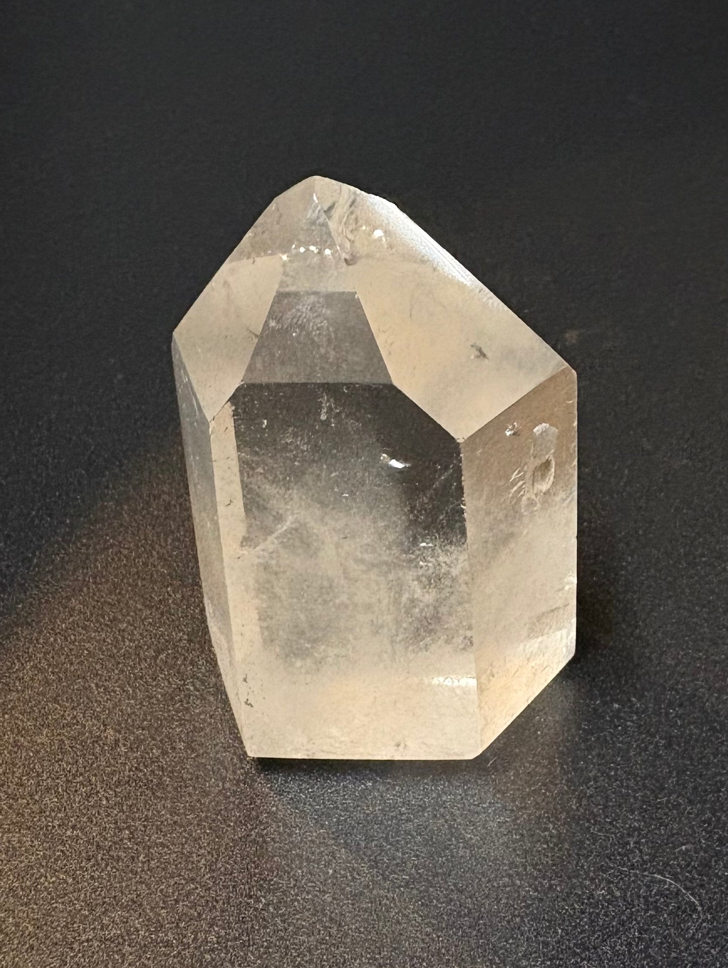 A natural clear quartz crystal point with sharp geometric facets, standing upright on a dark textured surface.