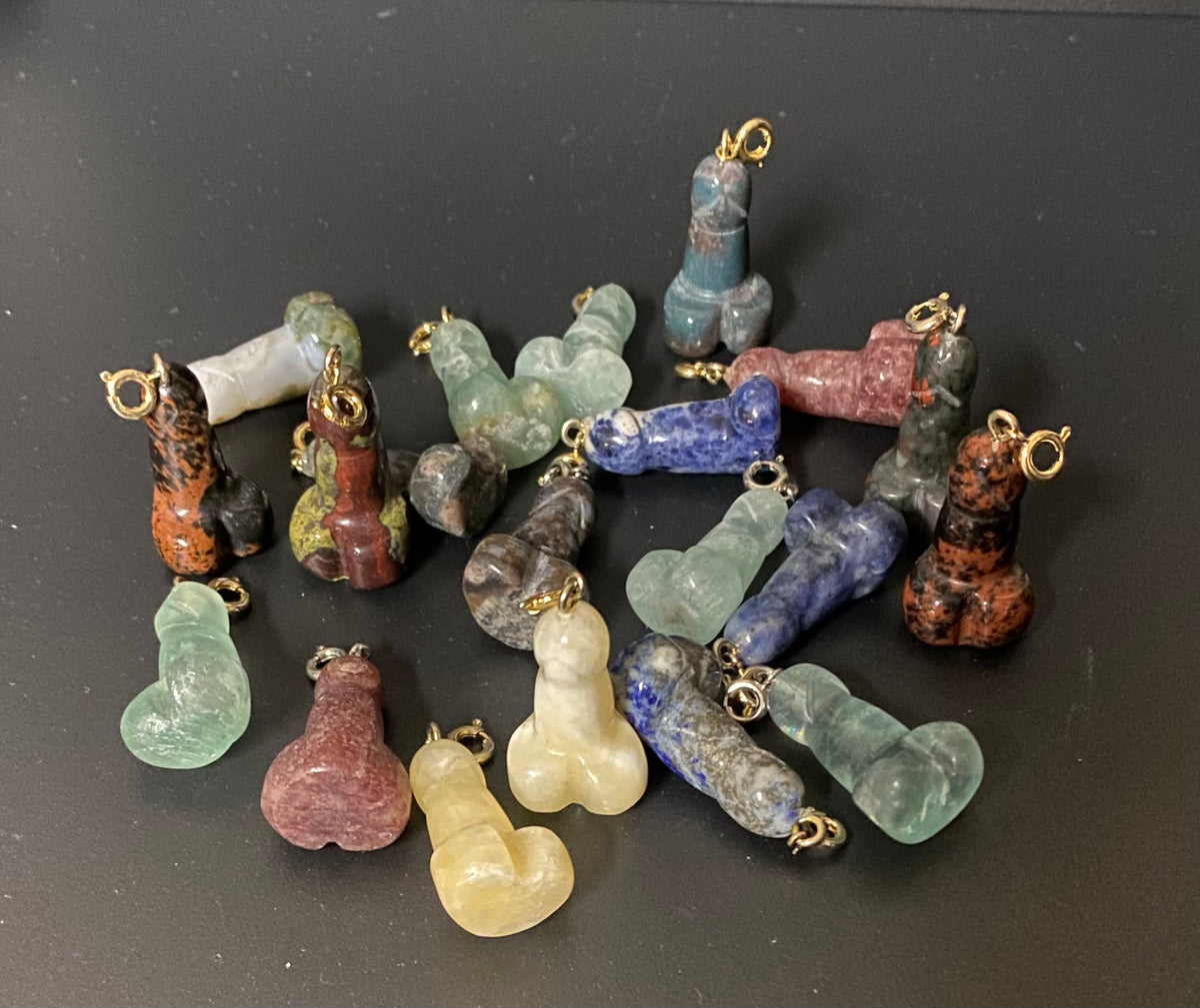 A group of small carved stone pendants shaped like male genitalia, each with a gold-tone loop at the top, arranged on a dark surface. The stones vary in color including blue, green, red, pink, yellow, brown, and gray.