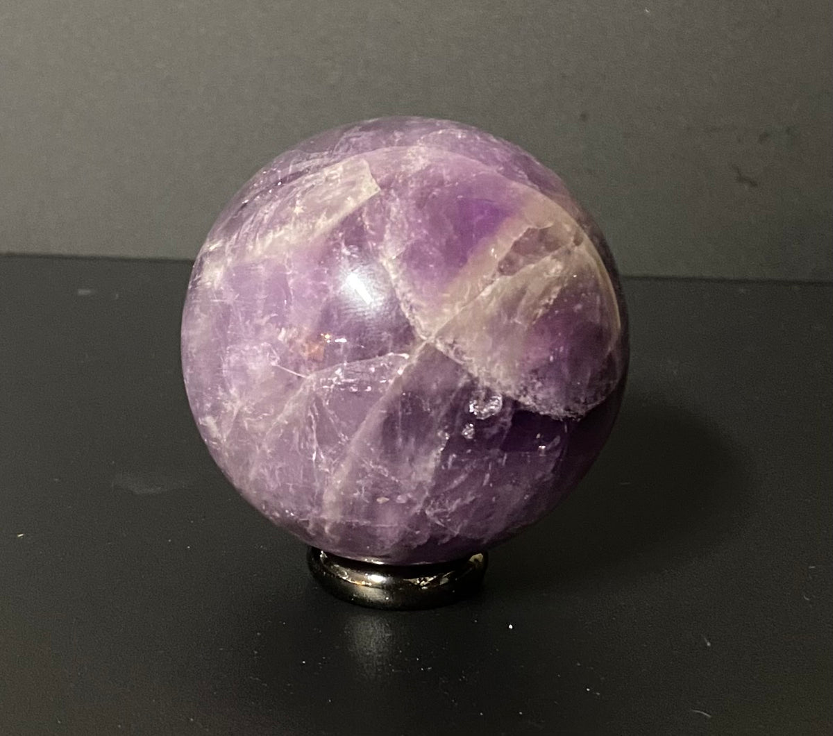 A polished purple amethyst crystal sphere resting on a small black metal stand against a dark gray background.