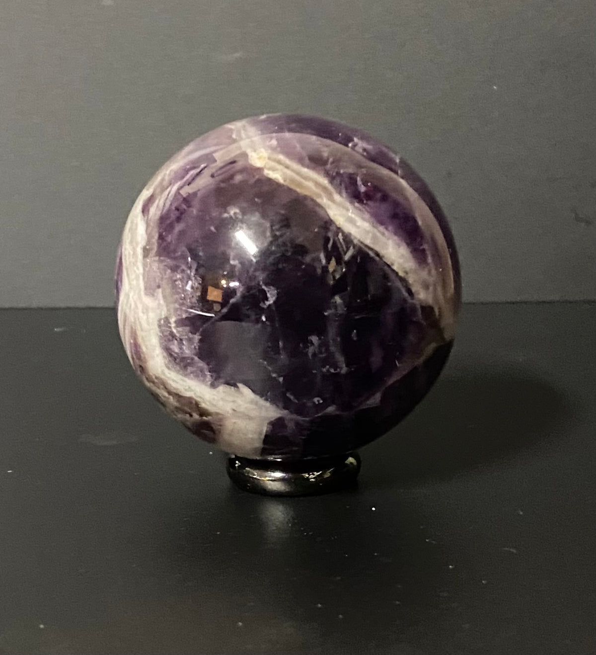 A polished purple amethyst crystal sphere with white veining, displayed on a small black stand against a dark gray background.