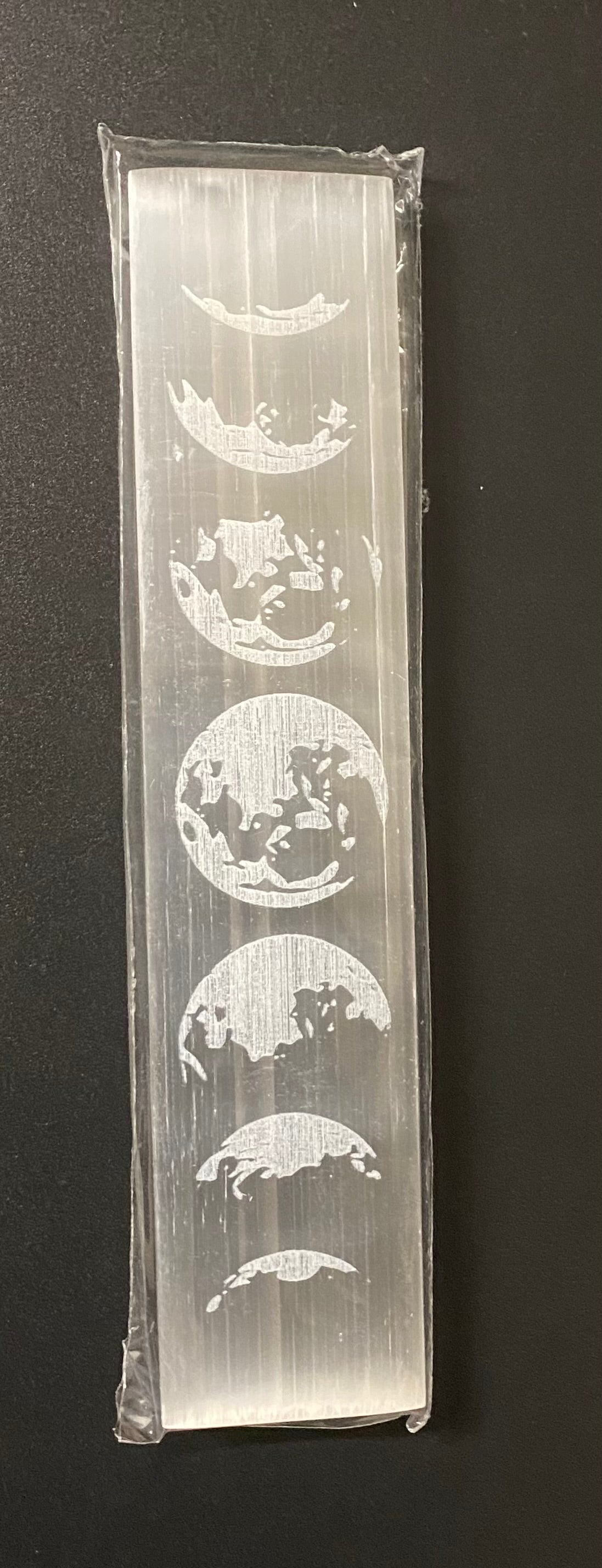 A selenite block featuring a vertical sequence of moon phases, sealed in a transparent plastic sleeve and photographed against a dark background.