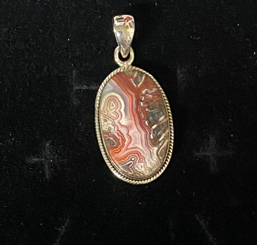An oval pendant featuring a polished agate stone with swirling bands of red, orange, white, and brown, set in a sterling silver bezel with a bail at the top, displayed on a black background.