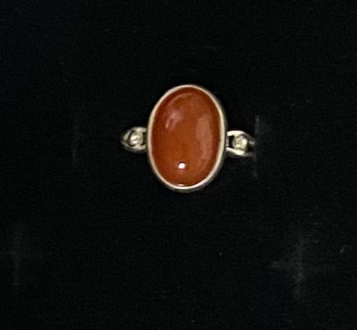 An oval ring featuring a polished reddish-brown cabochon gemstone in a silver-toned setting, flanked by two small round clear stones on a black background.
