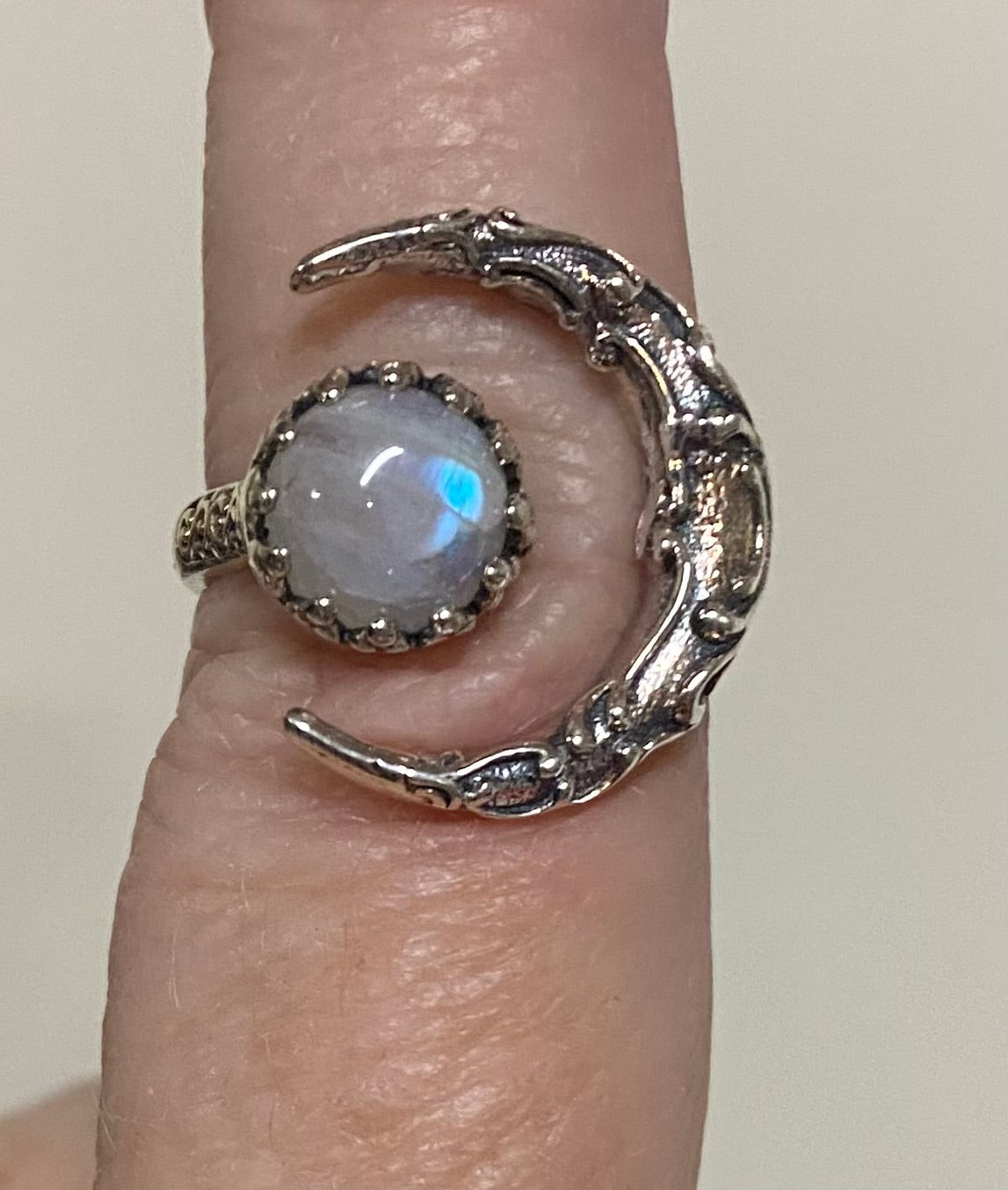 A close-up of a silver crescent moon ring with a round, iridescent moonstone cabochon, worn on a finger.
