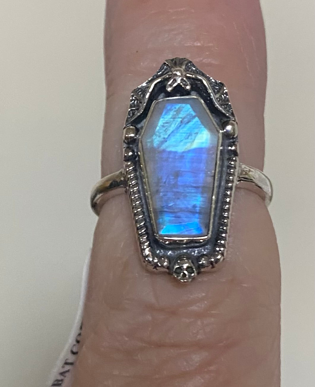 A silver ring with a coffin-shaped iridescent blue gemstone, shown worn on a finger.