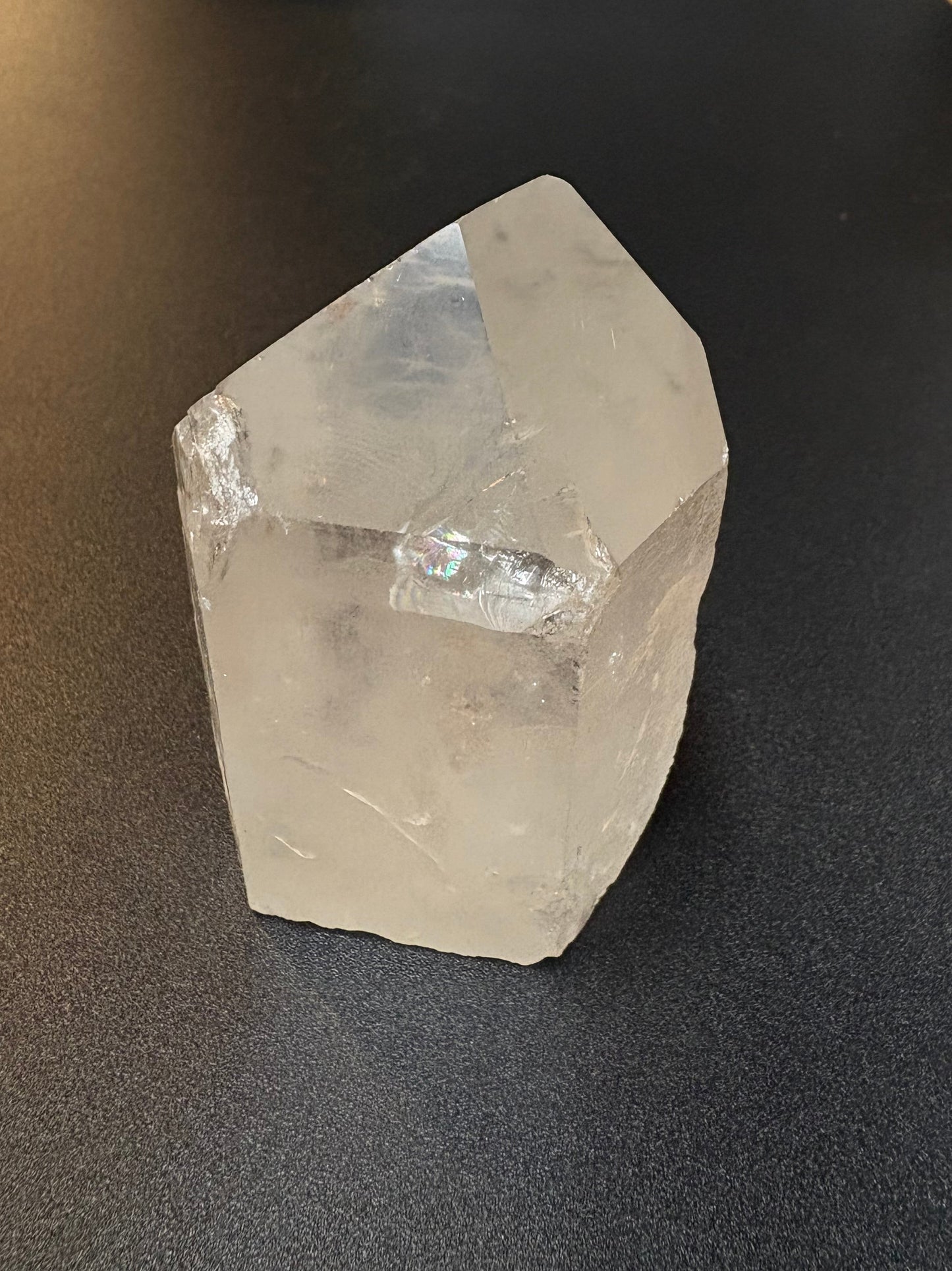 A raw, unpolished clear quartz crystal with visible natural facets and inclusions, resting on a dark textured surface.