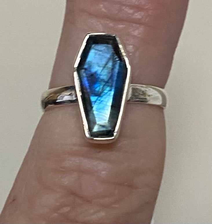 A silver ring featuring a coffin-shaped labradorite gemstone with a vibrant blue and green flash, worn on a finger.