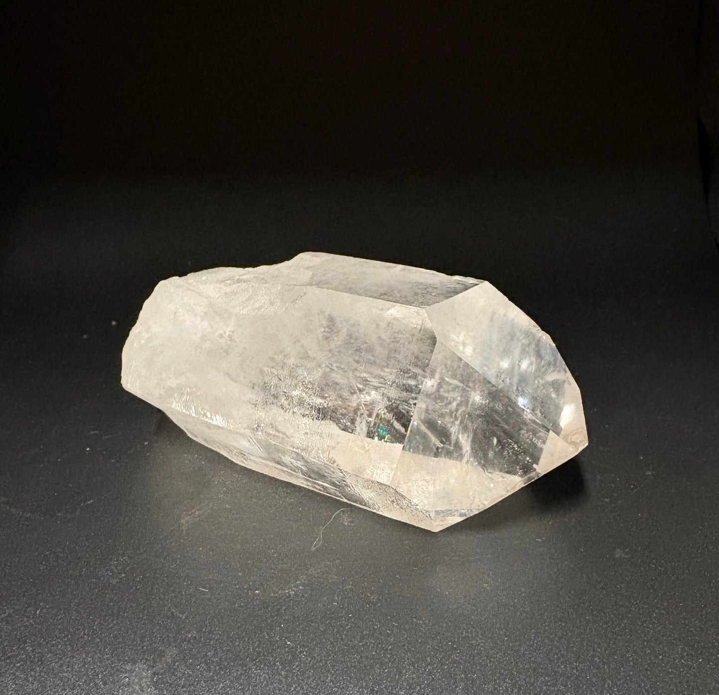 A raw, unpolished clear quartz crystal point resting on a dark gray surface against a black background.