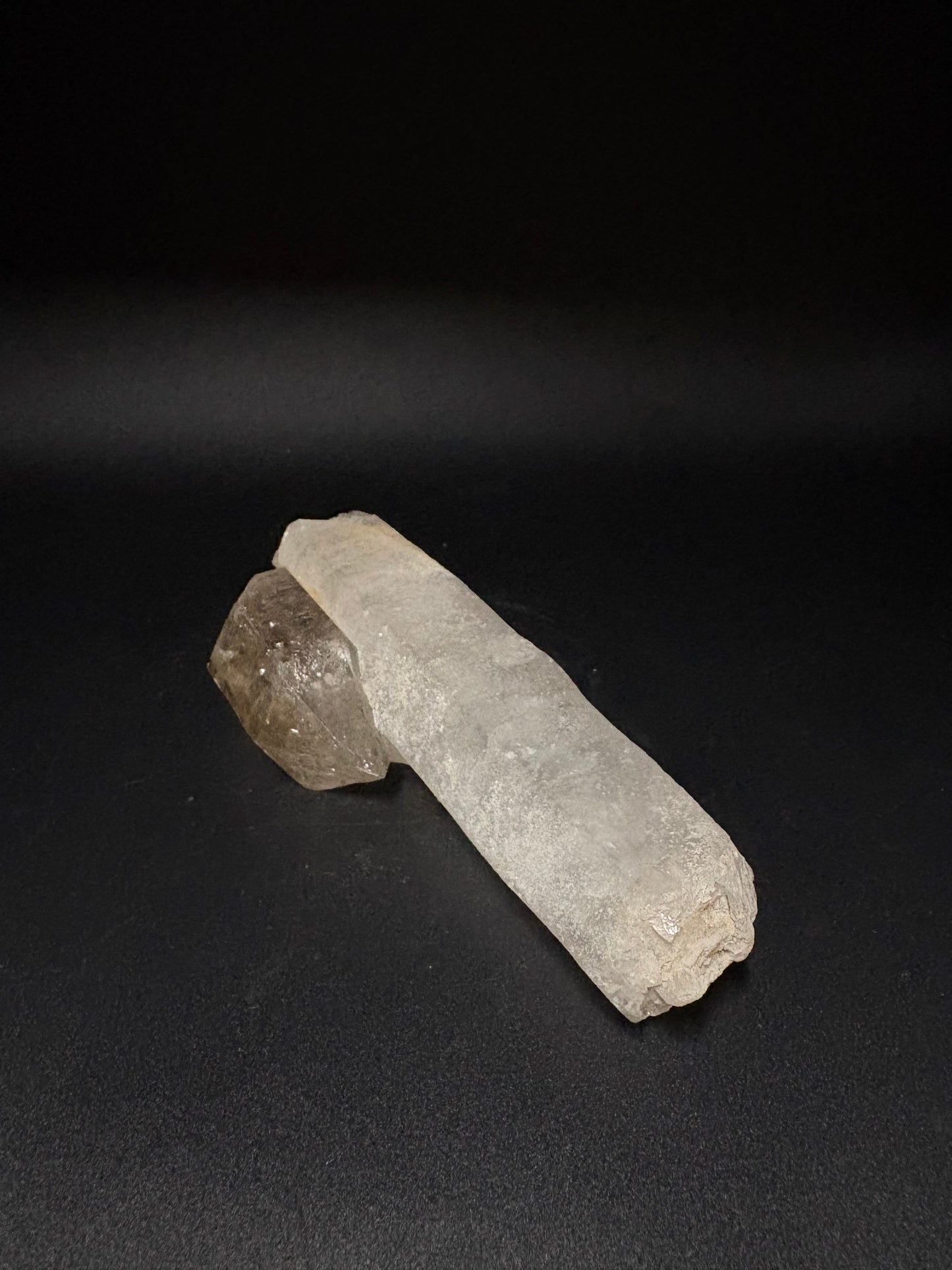 A raw crystal cluster featuring a long, pale gray translucent column and a smaller, rougher smoky brown piece, displayed on a dark black background.
