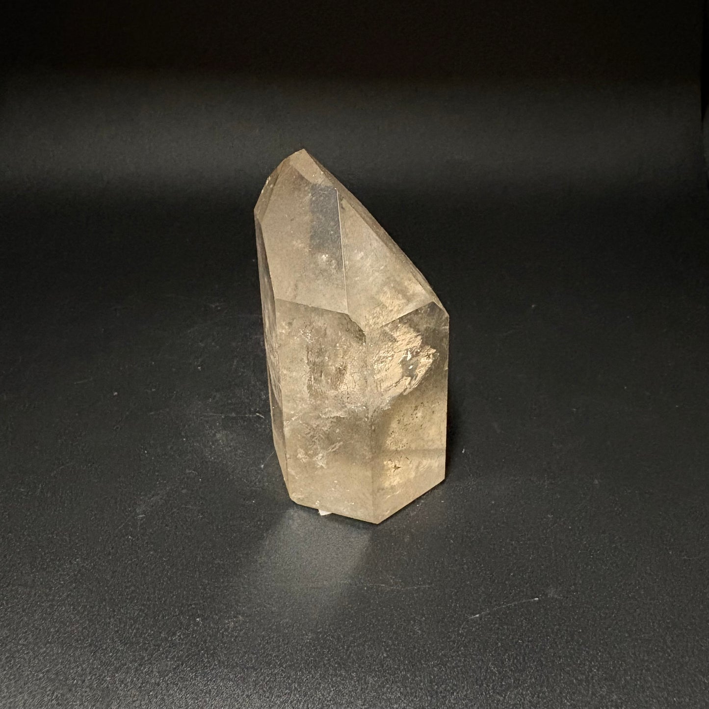 A natural translucent quartz crystal point with a warm beige-tan hue, standing upright on a dark gray surface against a dark background.