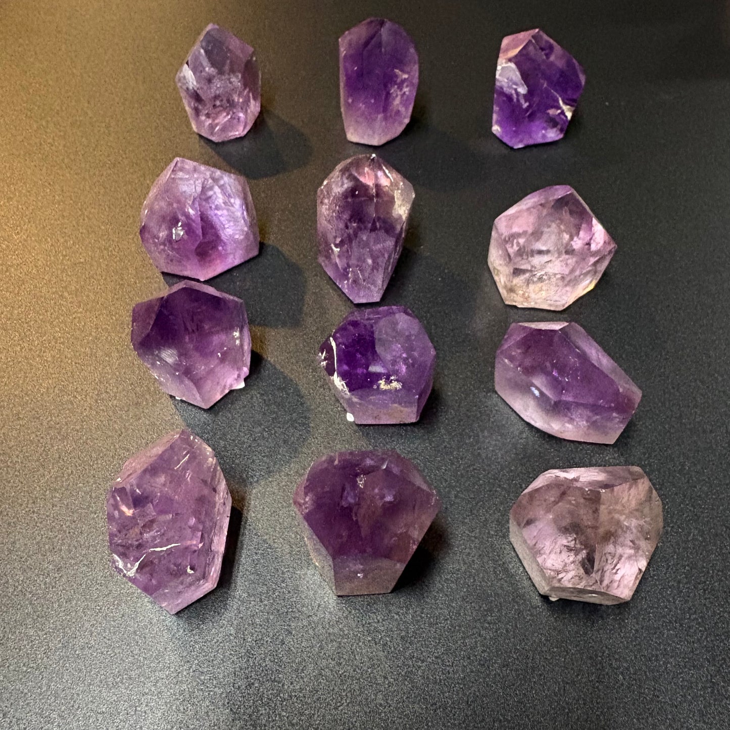 Twelve raw amethyst crystal points of various sizes arranged in a grid on a dark gray textured surface.
