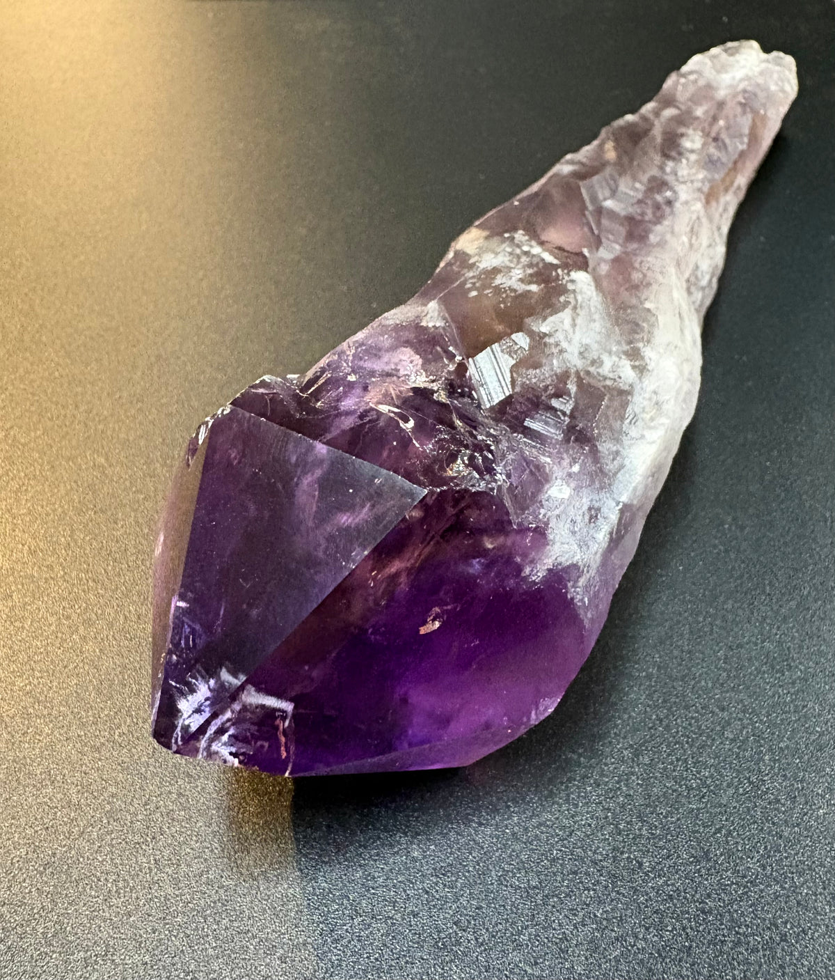 A raw amethyst crystal cluster with a natural pointed tip, showing a gradient from deep purple to milky white, resting on a dark gray surface.