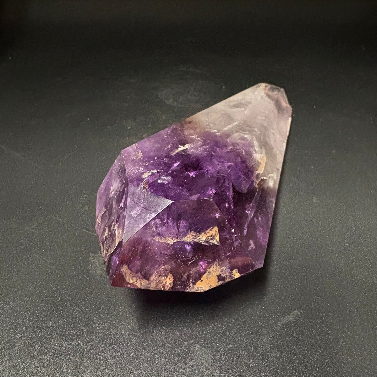A raw amethyst crystal point with deep purple and lighter lavender sections, resting on a dark gray textured surface.