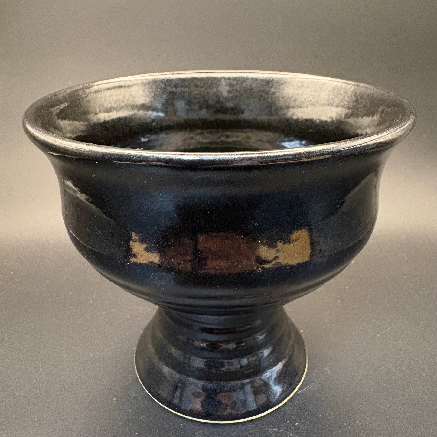 A dark glazed ceramic footed bowl with a glossy black finish and subtle brown and gold mottled accents, set against a neutral gray background.