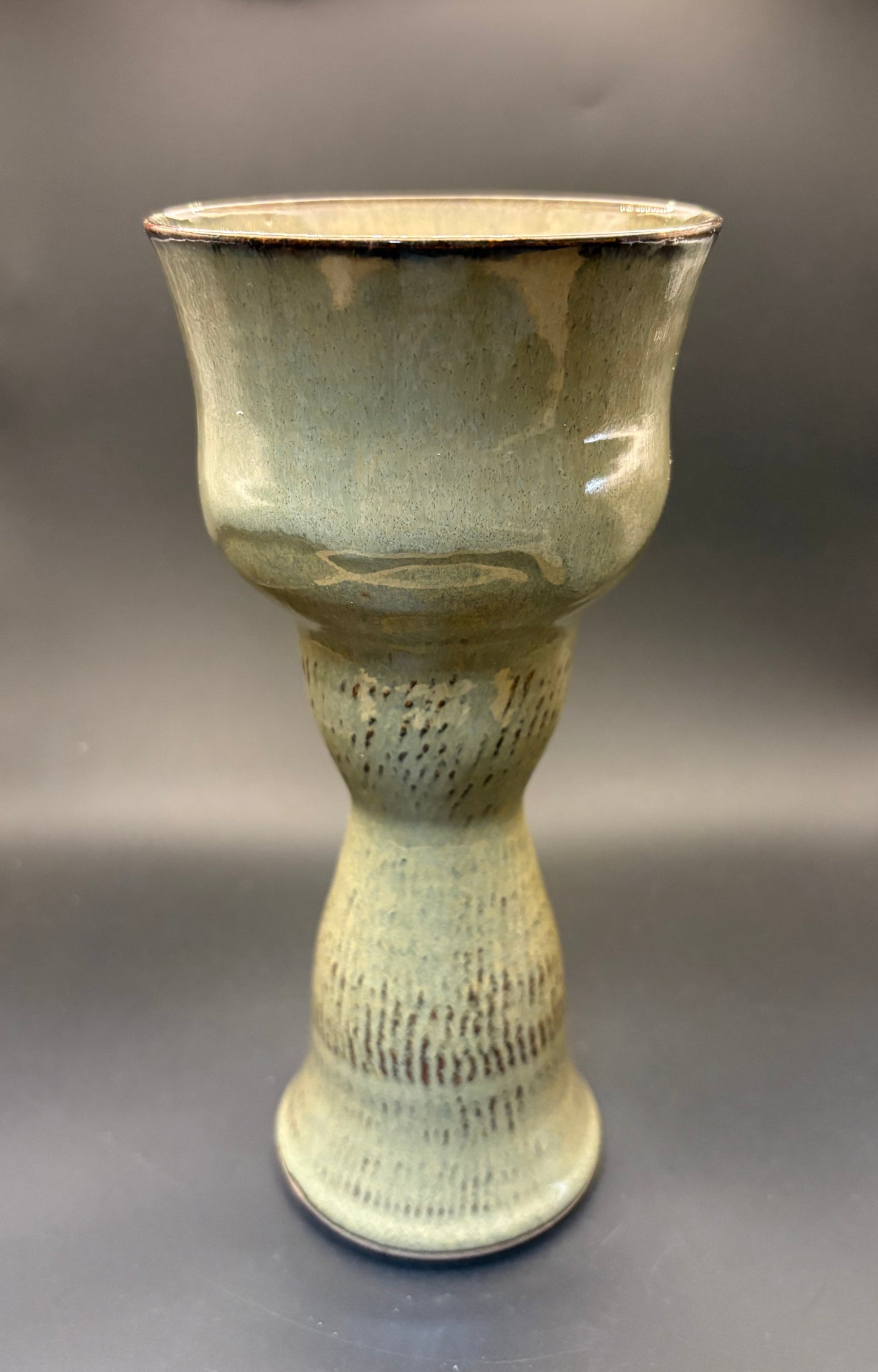 A handmade ceramic goblet or chalice with a textured, ribbed foot and a wide, flared rim, covered in a glossy sage green glaze with dark brown speckled accents, set against a dark gray background.