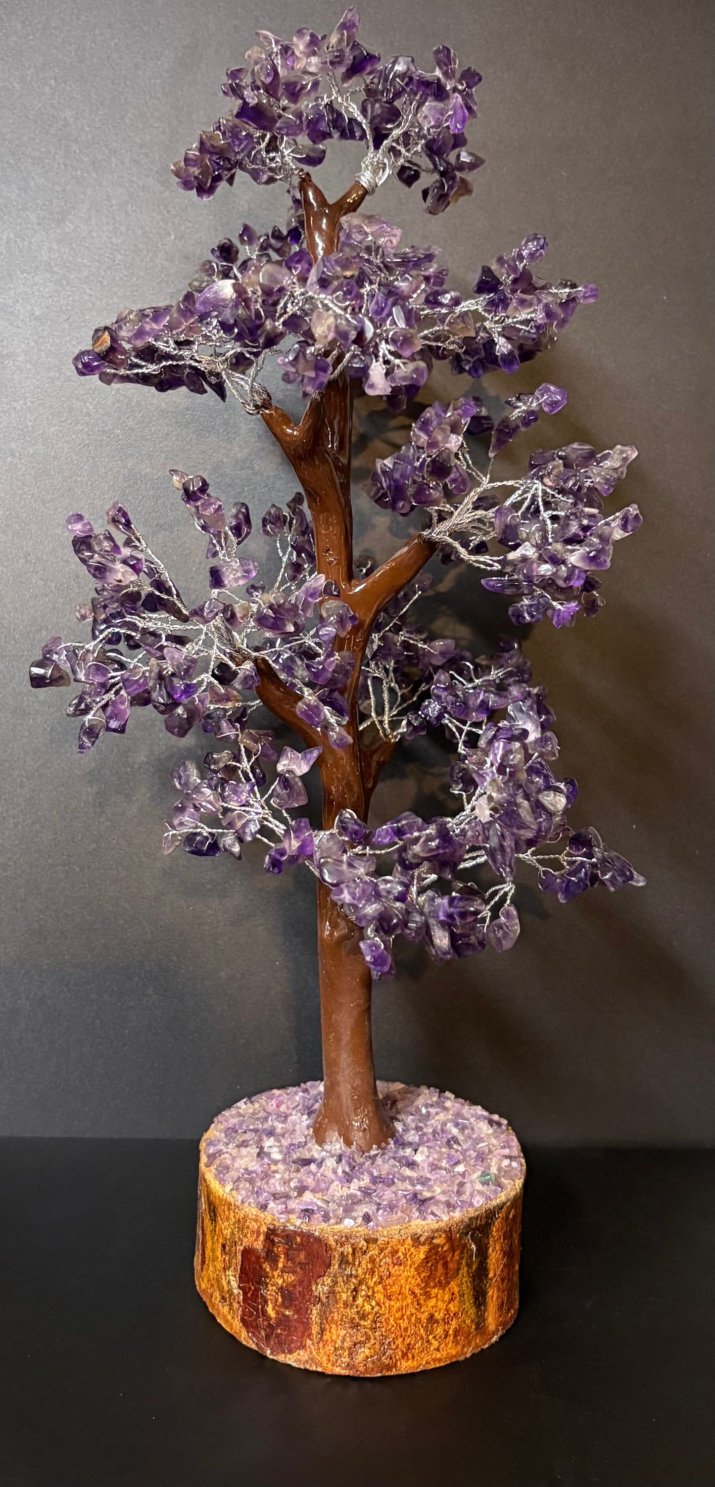 A decorative crystal tree sculpture with purple amethyst flower clusters on a brown branch structure, mounted on a round orange-brown stone base against a dark gray background.