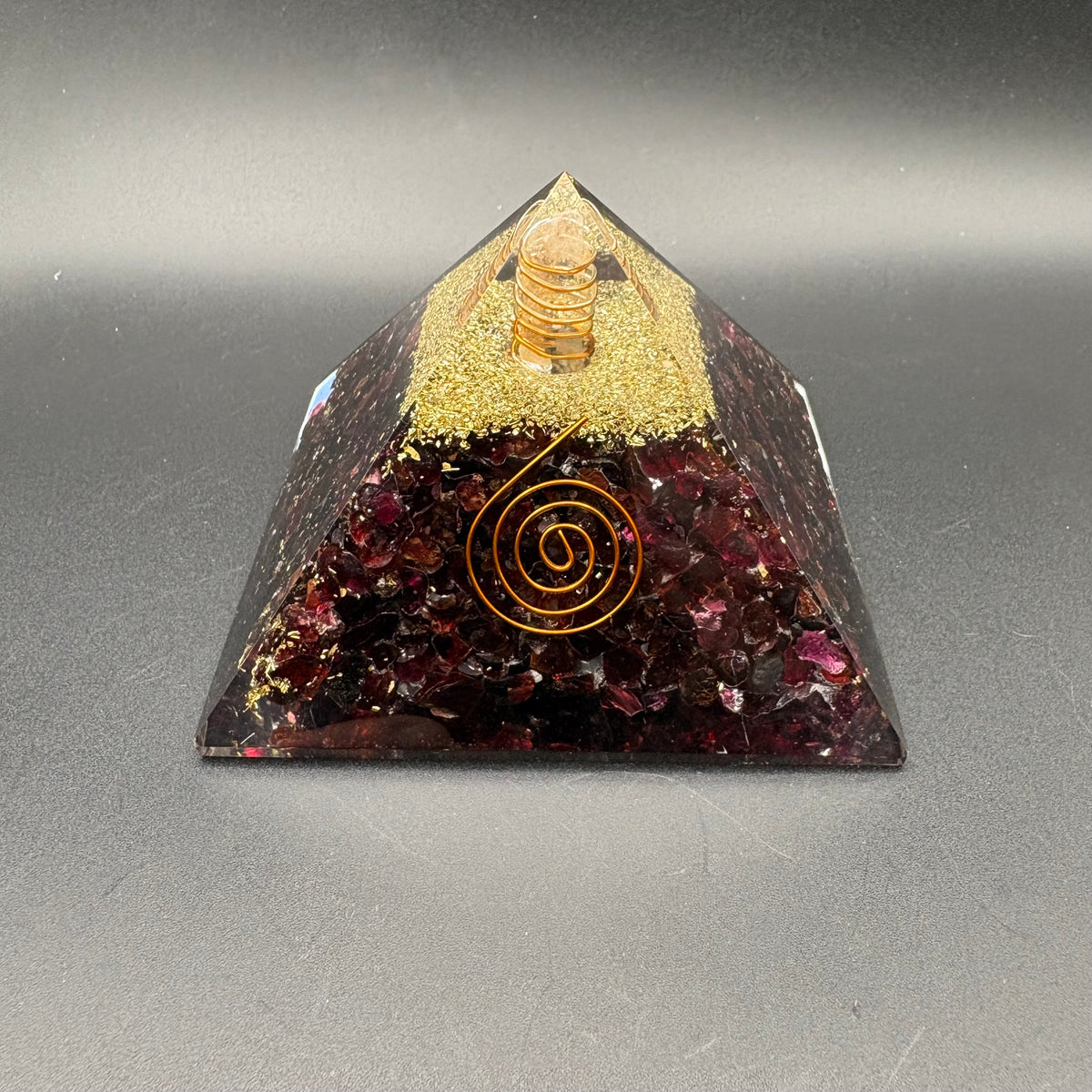 A triangular pyramid crystal tower with a gold spiral design on the front, filled with garnet stones, gold glitter, and a small clear quartz point at the top, set against a dark gray background.