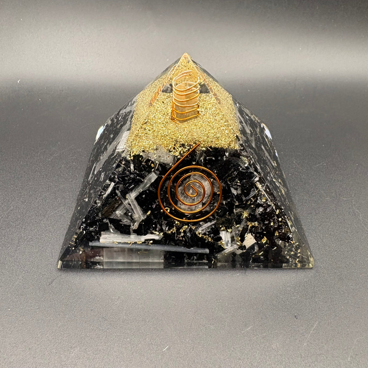 A clear resin pyramid with tourmaline and selenite inclusions, featuring a gold glitter top section, a gold spiral ornament, and a small copper coil at the apex, set against a neutral gray background.
