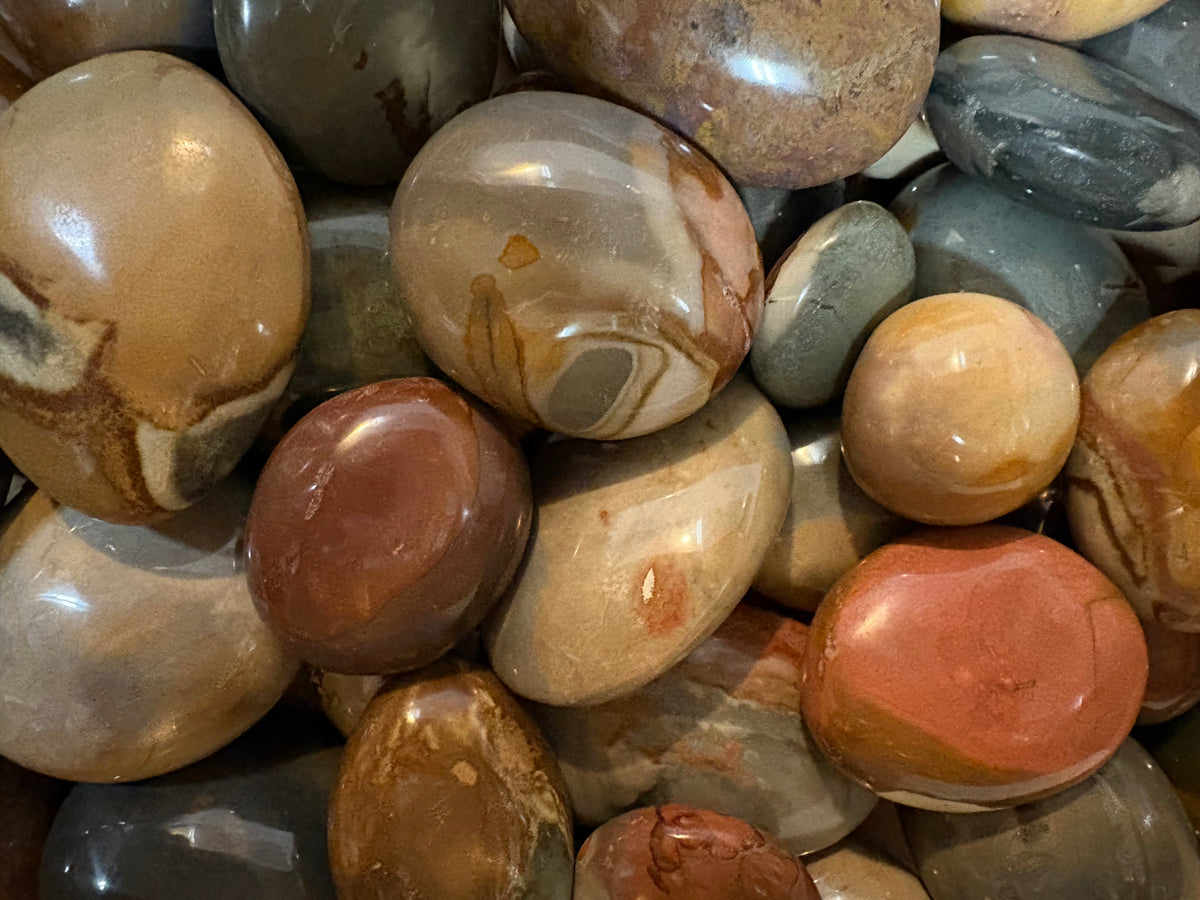 A close-up pile of polished Mookite stones ; earthy tones, including tan, brown, gray, muted red, and cream, with smooth rounded surfaces and visible natural banding.