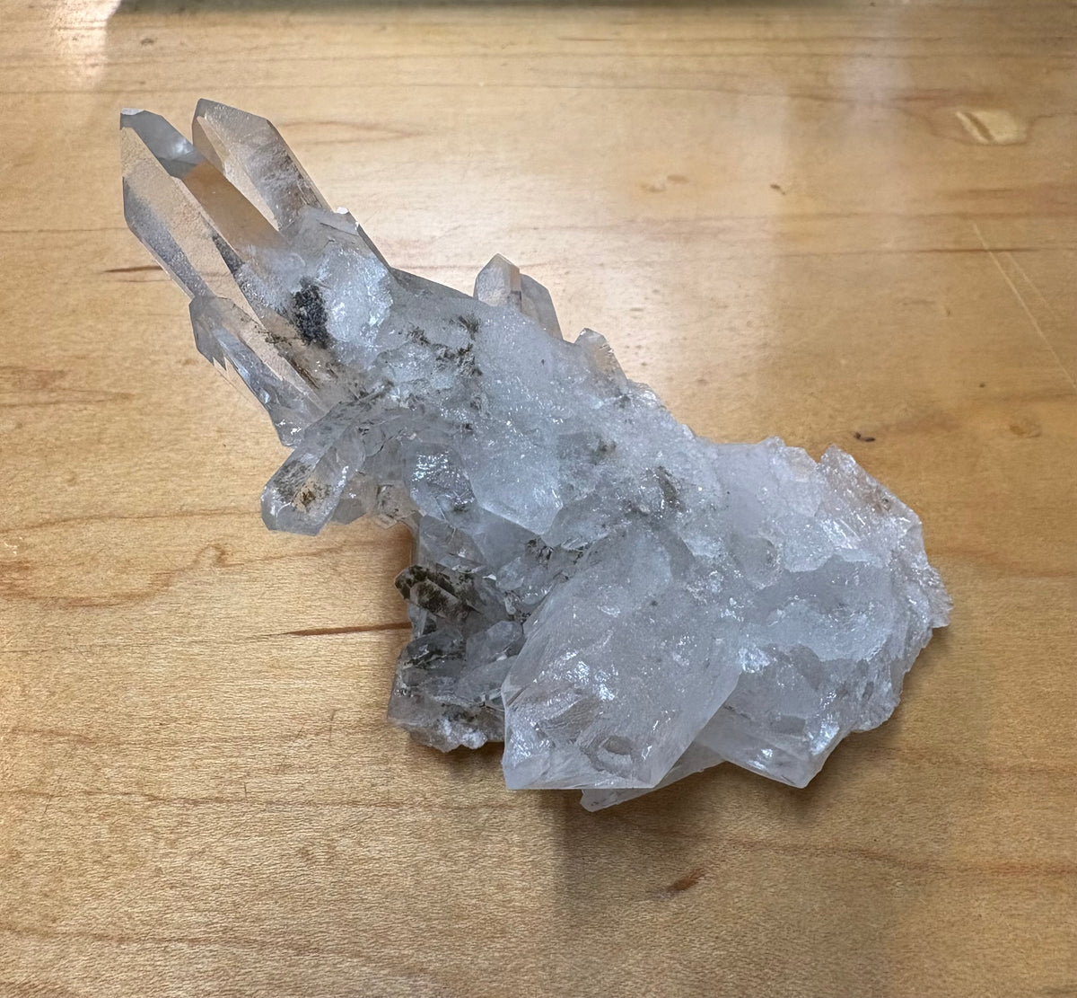 A natural clear quartz crystal cluster with multiple pointed tips and rough, textured facets, resting on a light brown wooden surface.