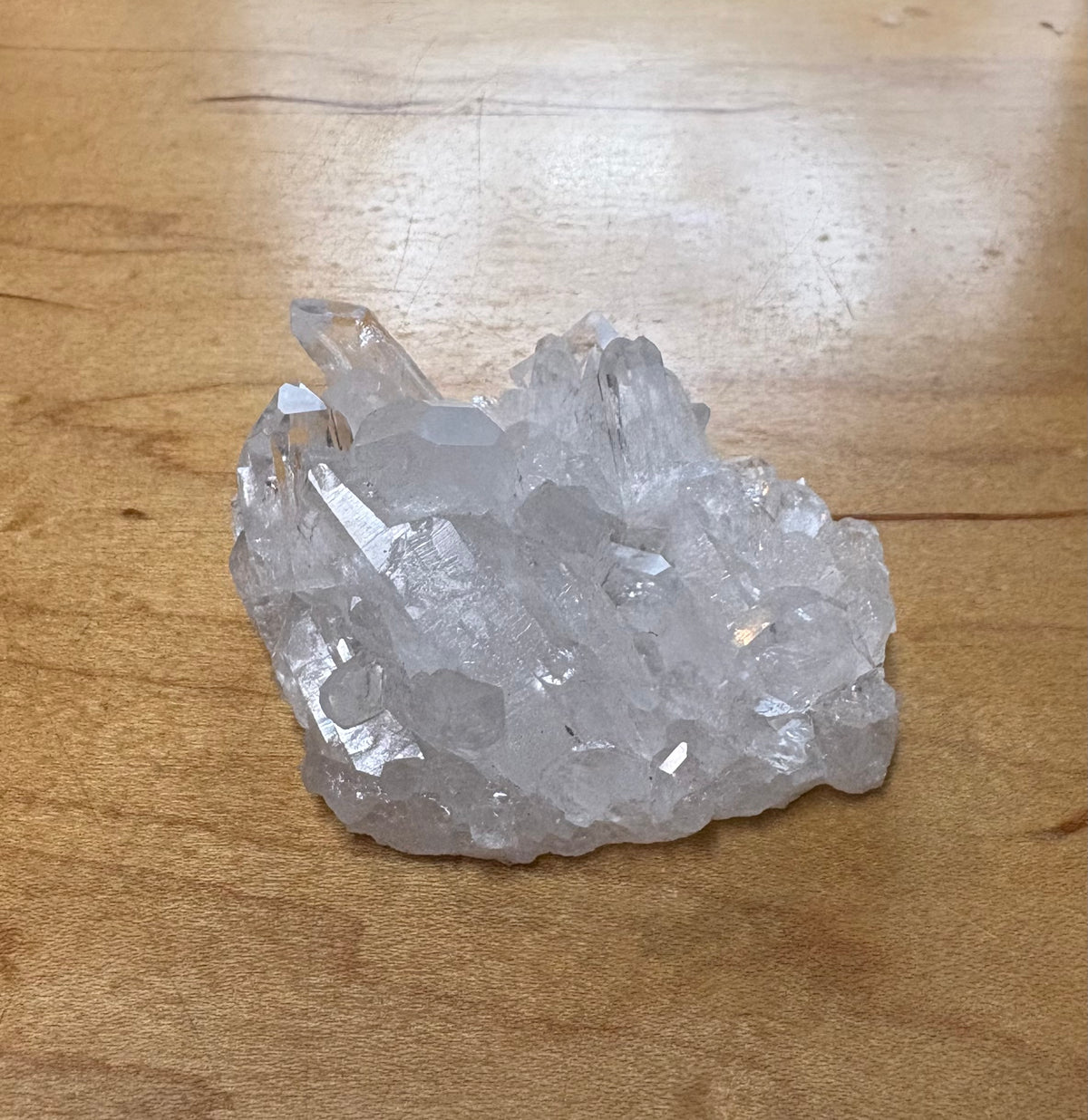 A raw crystal cluster with clear, faceted quartz-like points resting on a light brown wooden surface.