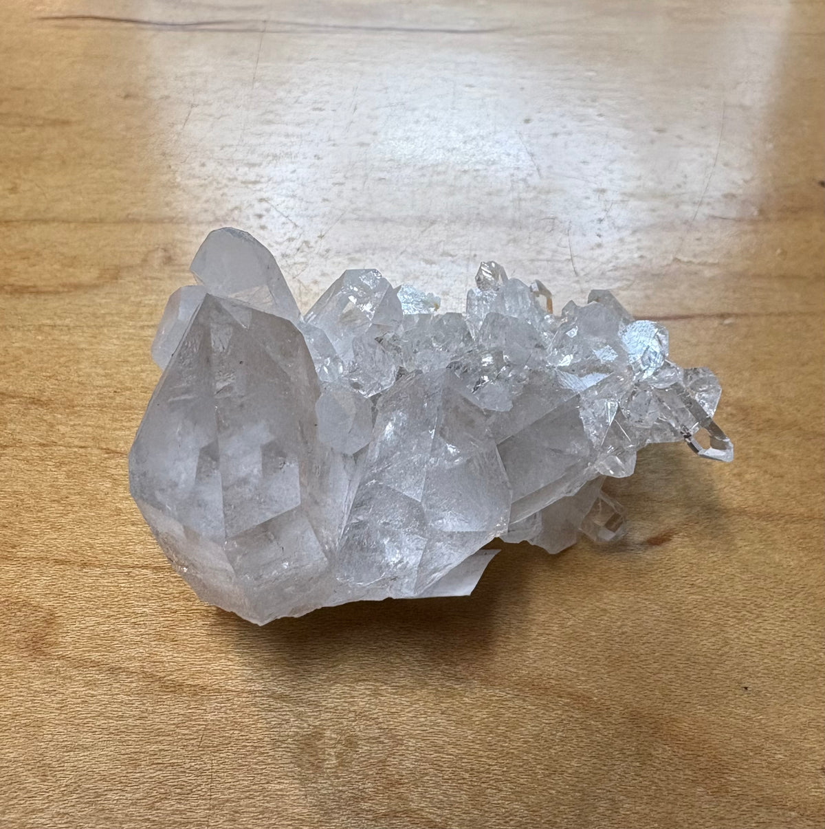 A natural clear quartz crystal cluster resting on a light brown wooden tabletop.
