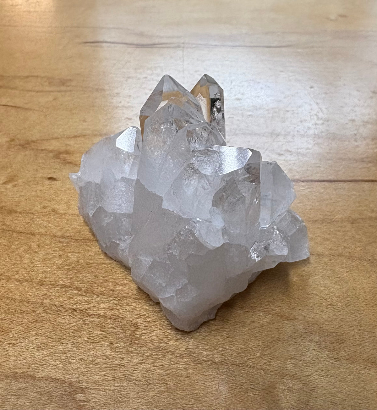 A raw crystal cluster with multiple translucent clear crystal points resting on a light brown wooden surface.