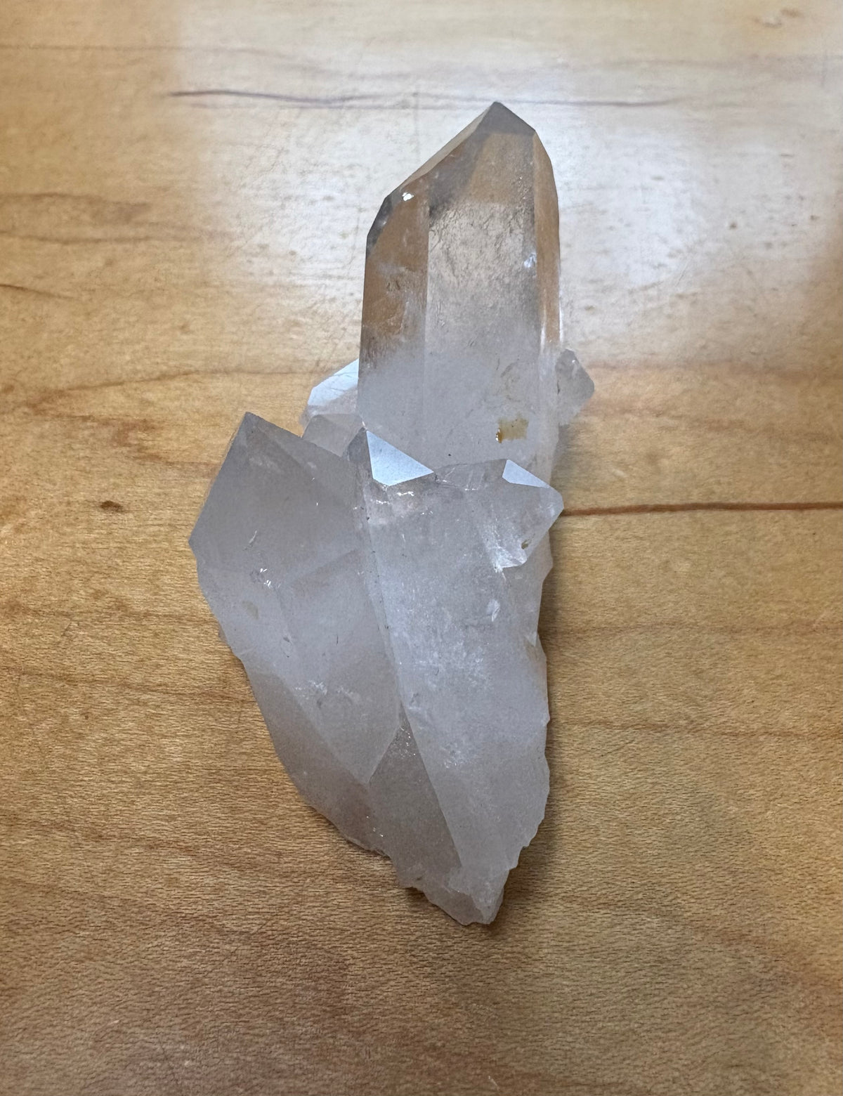 A raw clear quartz crystal cluster with a prominent central point resting on a light brown wooden surface.
