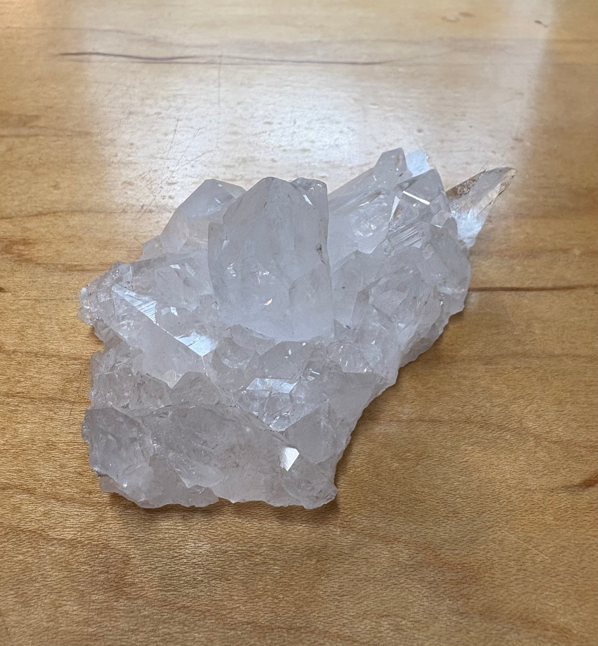 A raw crystal cluster, likely quartz, with multiple clear faceted points, resting on a light brown wooden surface.