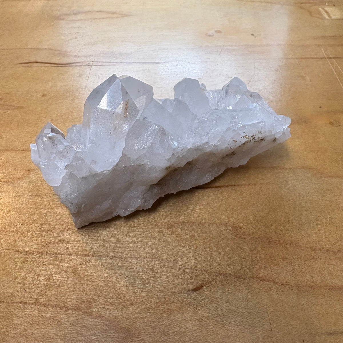 A raw crystal cluster with clear, faceted quartz points resting on a light brown wooden surface.