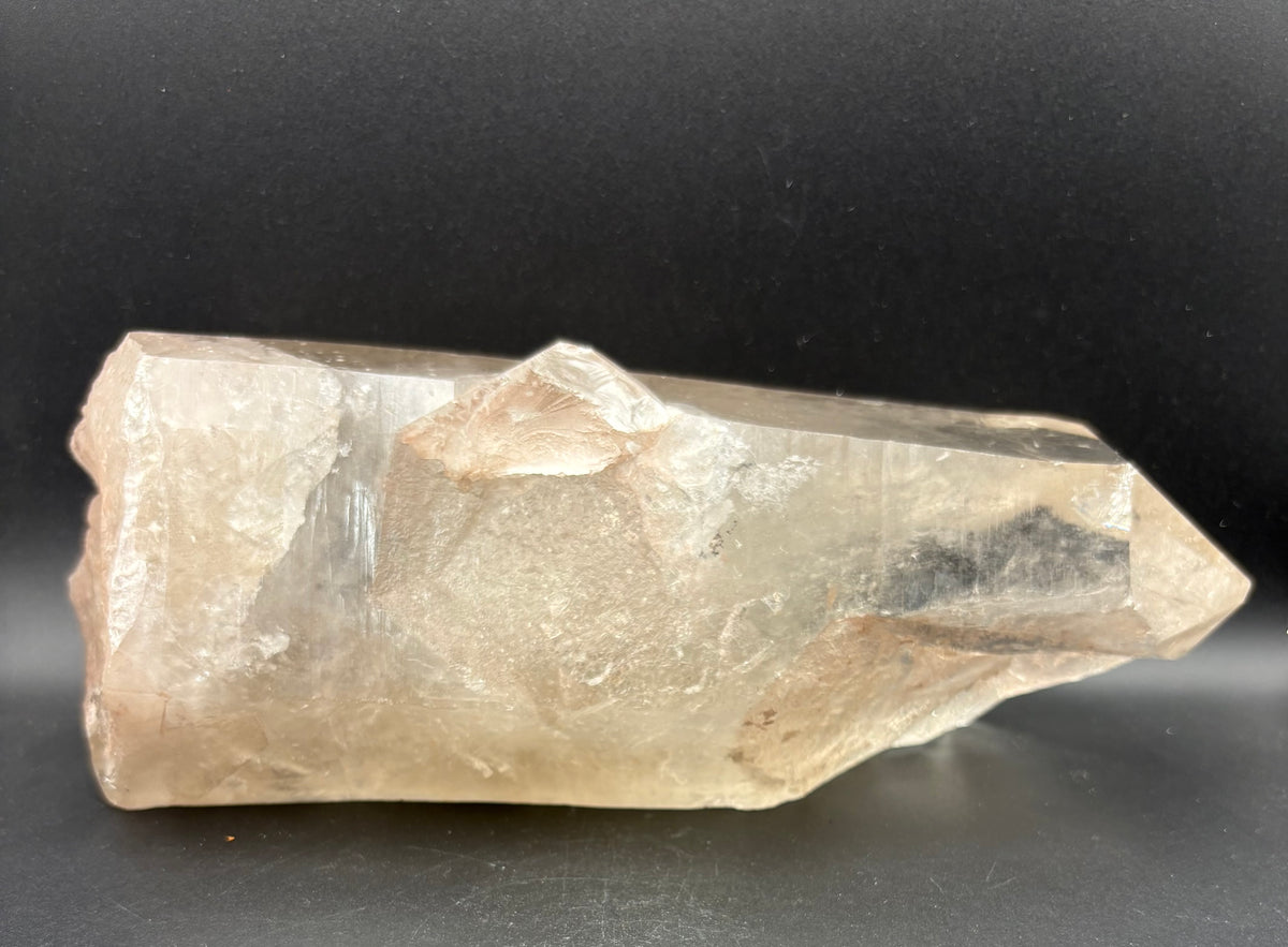 A raw, unpolished smoky quartz crystal point with natural inclusions and a warm beige-tan hue, resting on a dark black surface.
